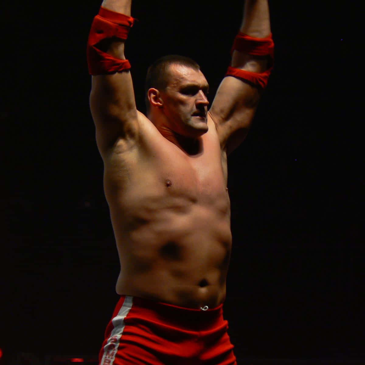 Buff Bagwell WWE舉手照片 壁紙 — 一個肌肉發達的摔角手，身穿紅色短褲和護腕，在黑暗背景下勝利地高舉雙臂。