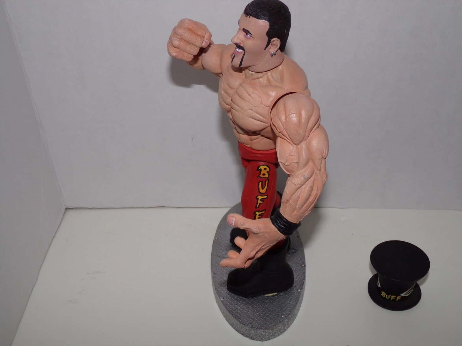 Buff Bagwell 拳擊手動作玩偶揮拳照片 壁紙 — 一個肌肉發達的摔角手人偶，穿著印有「BUFF」字樣的紅色短褲，在灰色底座上擺姿勢，旁邊放著一頂相配的黑色高帽。