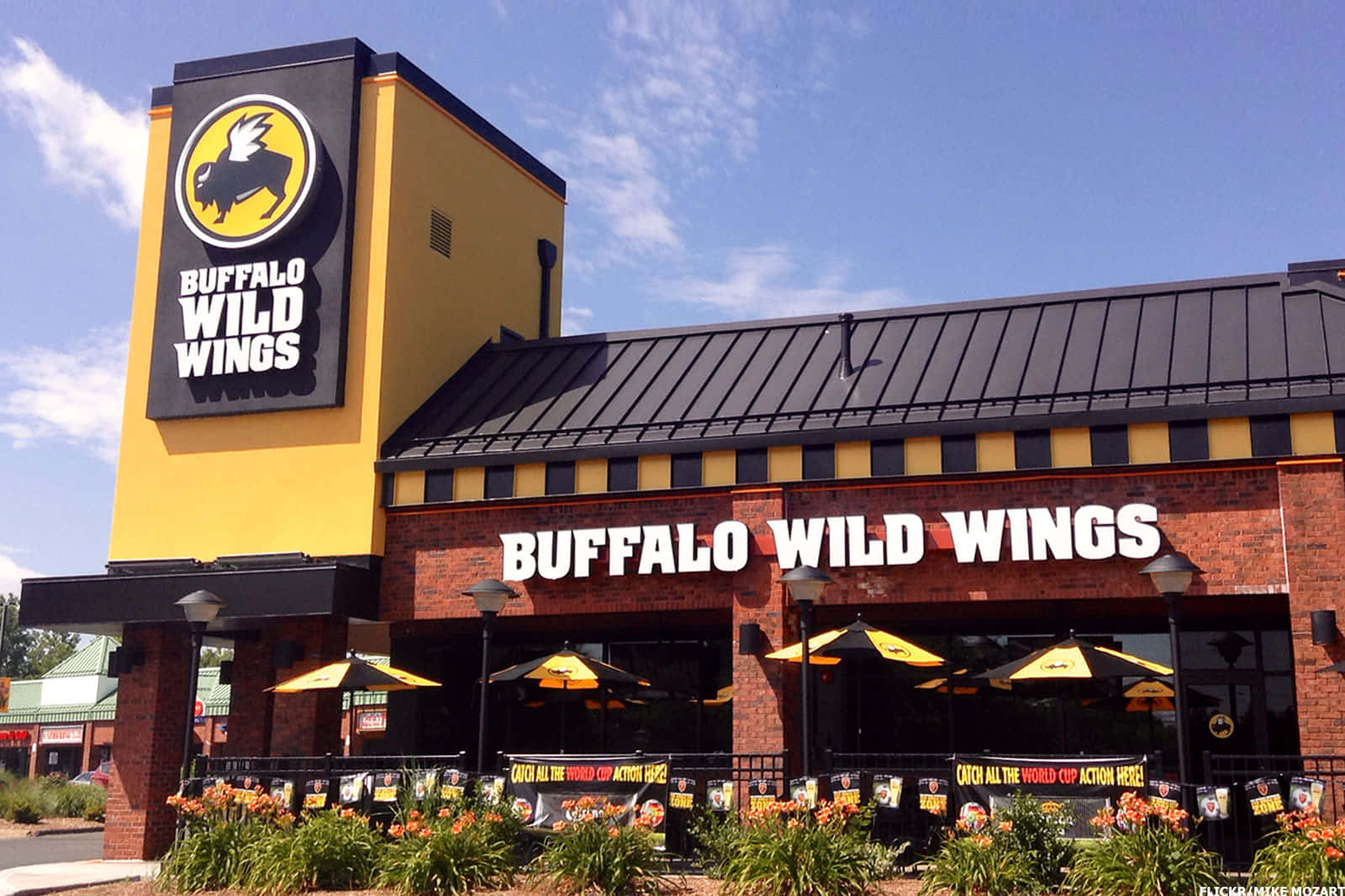 Buffalo Wild Wings的美味盛宴！ 壁紙 — 陽光普照的日子裡，一間明亮的黃色Buffalo Wild Wings餐廳，磚砌外牆，黃色太陽傘下設有戶外座位。