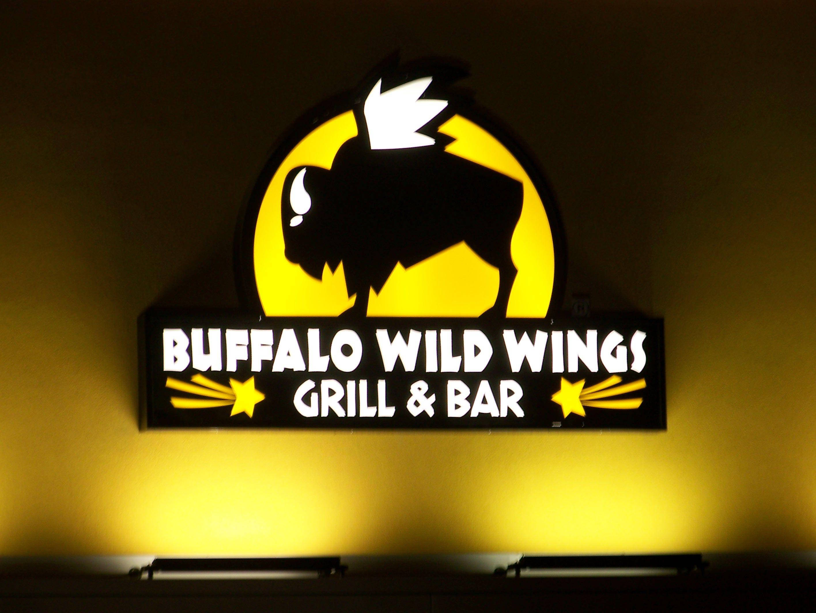 Buffalo Wild Wings 燈光招牌 壁紙 — 一個明亮照明的Buffalo Wild Wings標誌，黑色野牛剪影映襯在黃色太陽背景上，在牆上投射出溫暖的光芒。