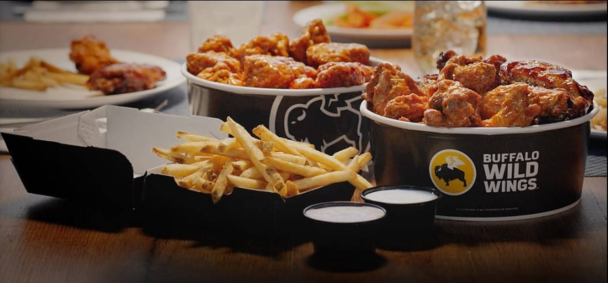 Buffalo Wild Wings 配薯條 壁紙 — 脆皮水牛城雞翼和金黃薯條盛載於品牌容器中，放在木桌上，附有蘸醬。
