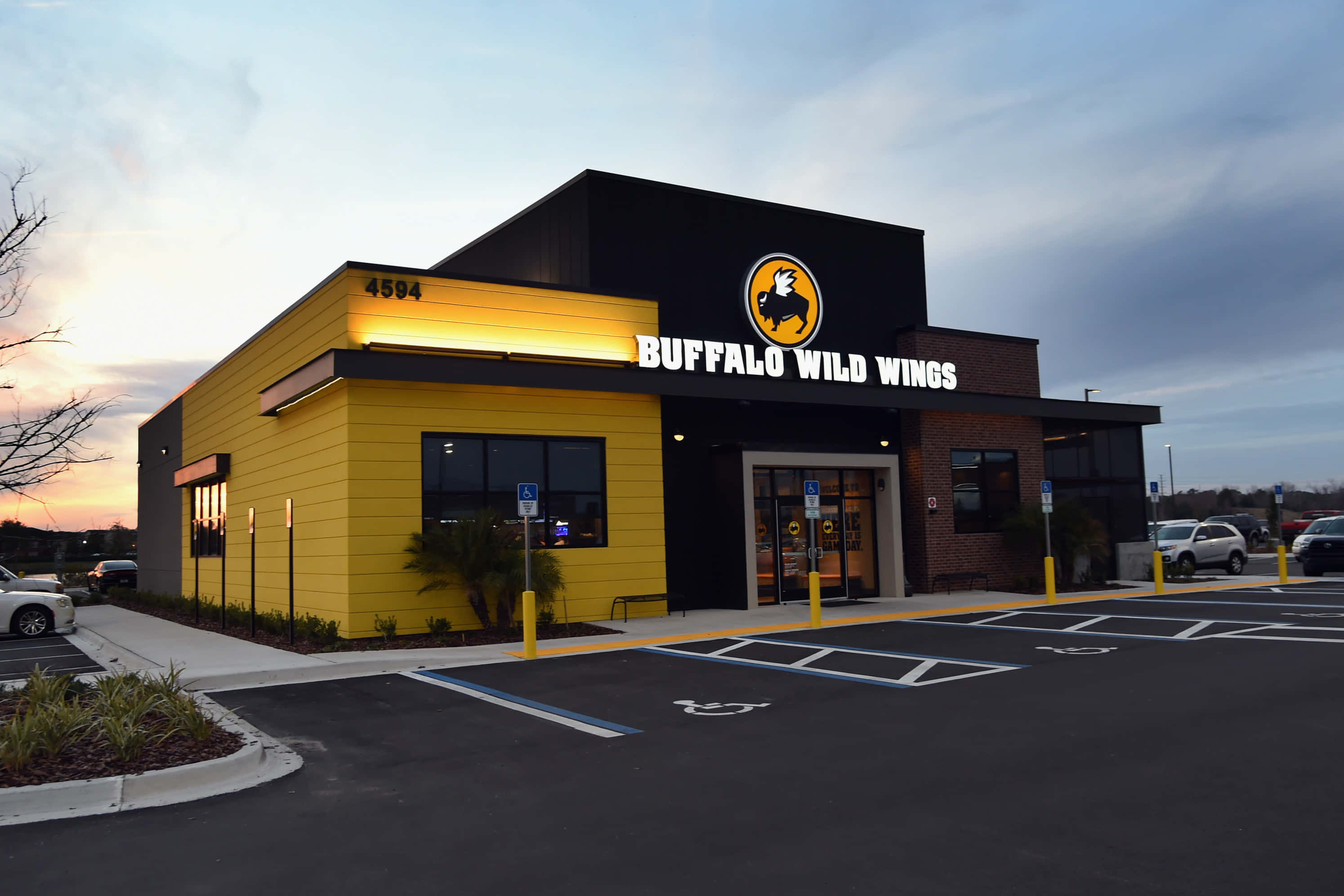 立即在Buffalo Wild Wings點購雞翅！ 壁紙 — 一間 Buffalo Wild Wings 餐廳在黃昏時分，鮮黃色牆壁和發光招牌在陰天天空下。