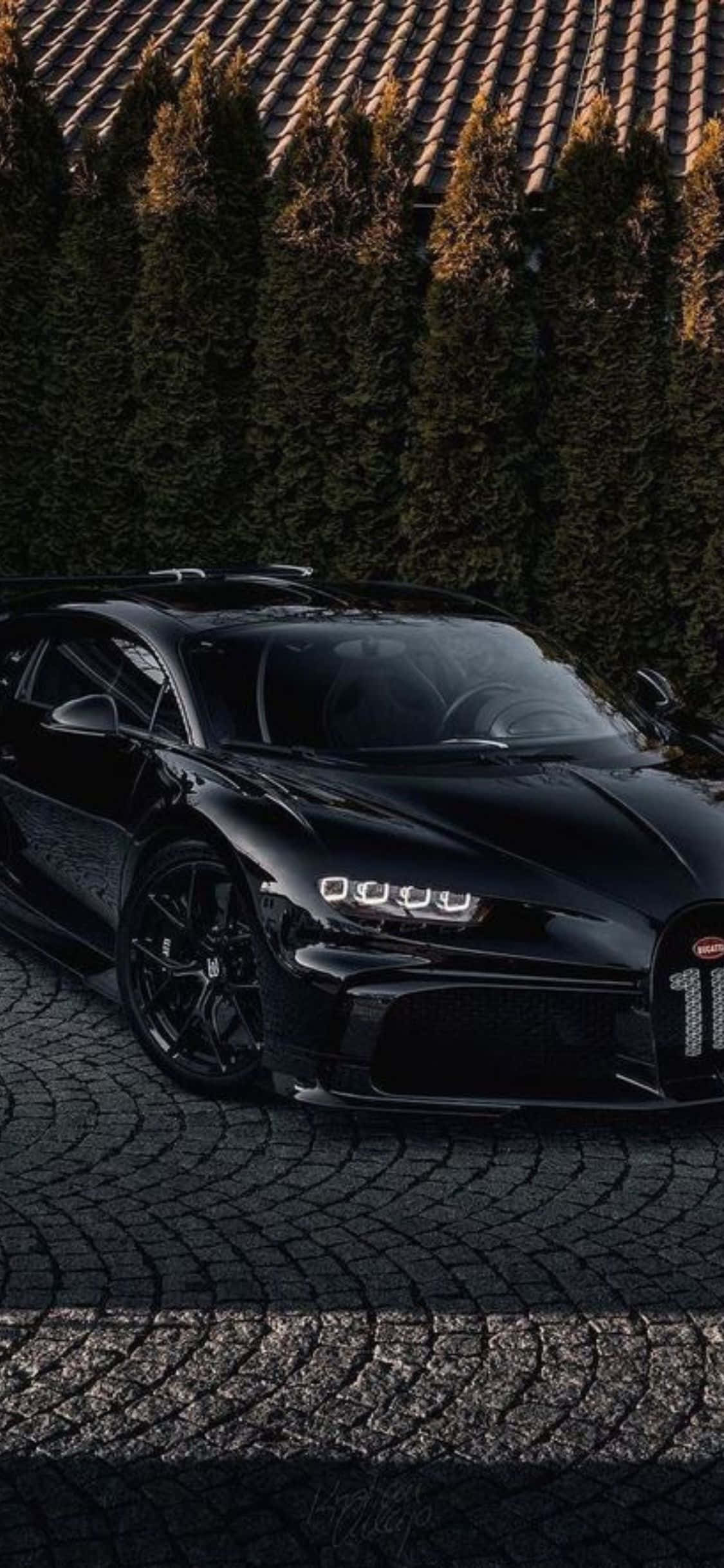 Bugatti Veyron 登上全新 iPhone XS 壁紙 — 一輛時尚的黑色Bugatti Chiron停泊在鵝卵石車道上，背後有高大的常綠樹，沐浴在溫暖的金色光線中。