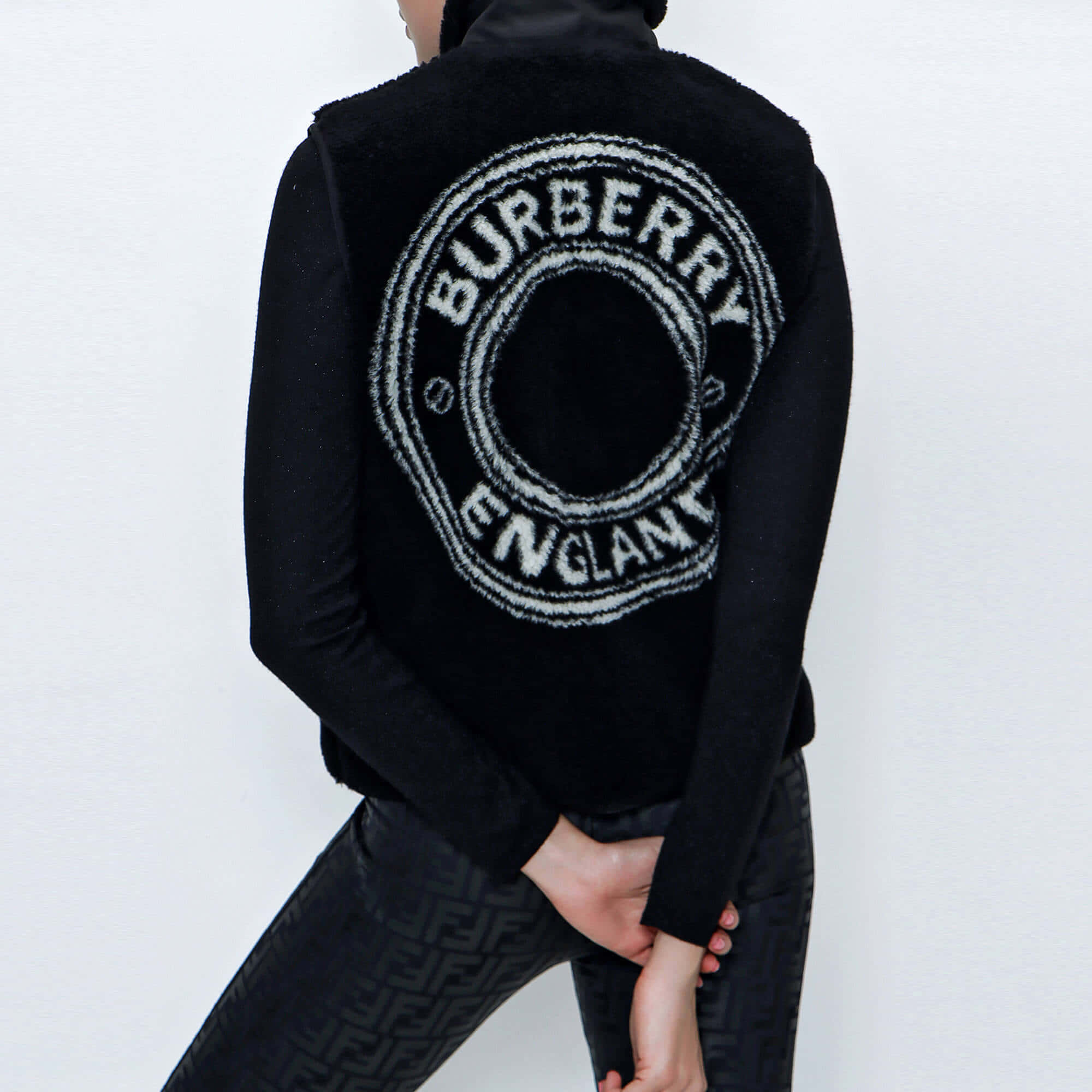 以Burberry展現時尚宣言 壁紙 — 背面視角，一個人穿著黑色Burberry背心，帶有白色圓形標誌，背景是純白牆壁。