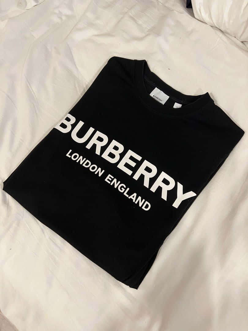 讓 Burberry 奢華時尚定義您的風格 壁紙 — 一件黑色Burberry T恤，上面有粗體白色字樣，整齊地折疊在皺巴巴的白色床單上。