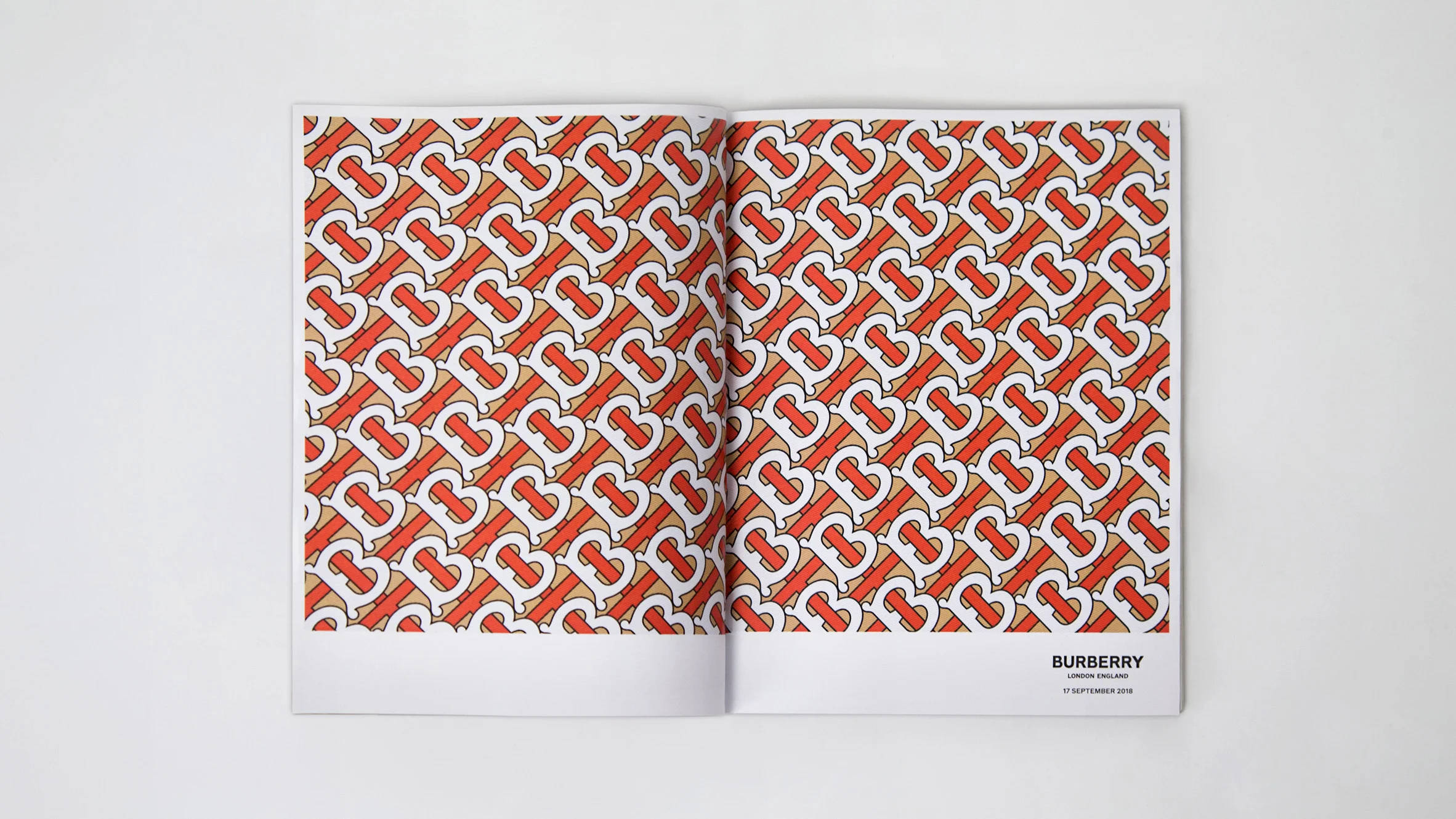 展示Burberry設計師標誌的雜誌內頁 壁紙 — 一本打開的雜誌在純白色背景上展示米色頁面上重複的紅白交錯「B」標誌大膽圖案。