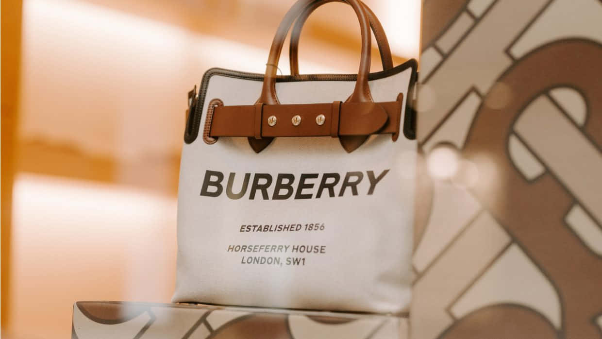 經典 Burberry 風格 壁紙 — 一個白色Burberry手袋配棕色皮革肩帶，優雅地放置在溫暖光線下的模糊圖案背景前。
