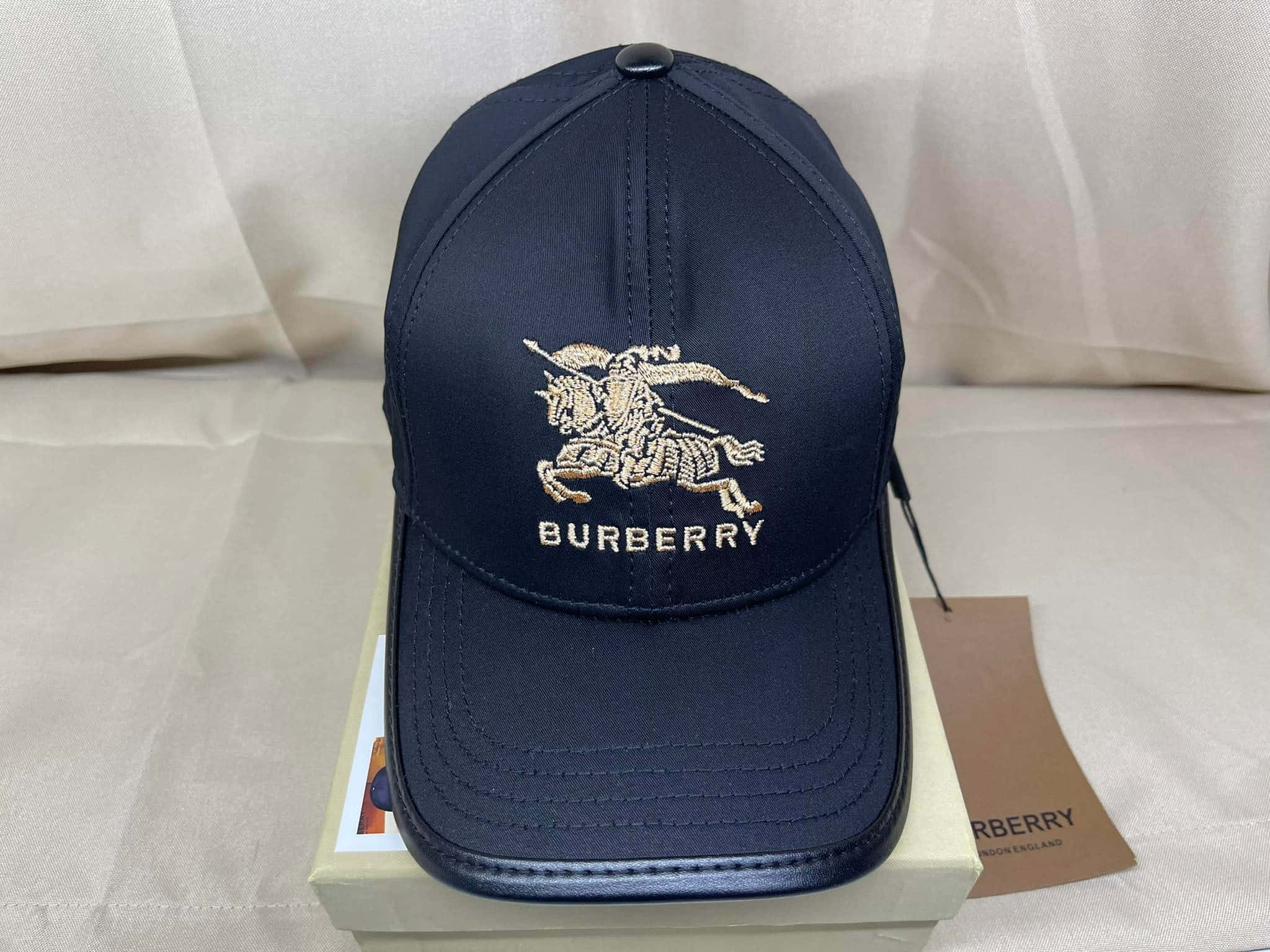 Burberry 最新系列奢華配件 壁紙 — 海軍藍色 Burberry 棒球帽，帶有金色刺繡騎士標誌，放在其品牌盒子上，背景為柔軟白色布料。
