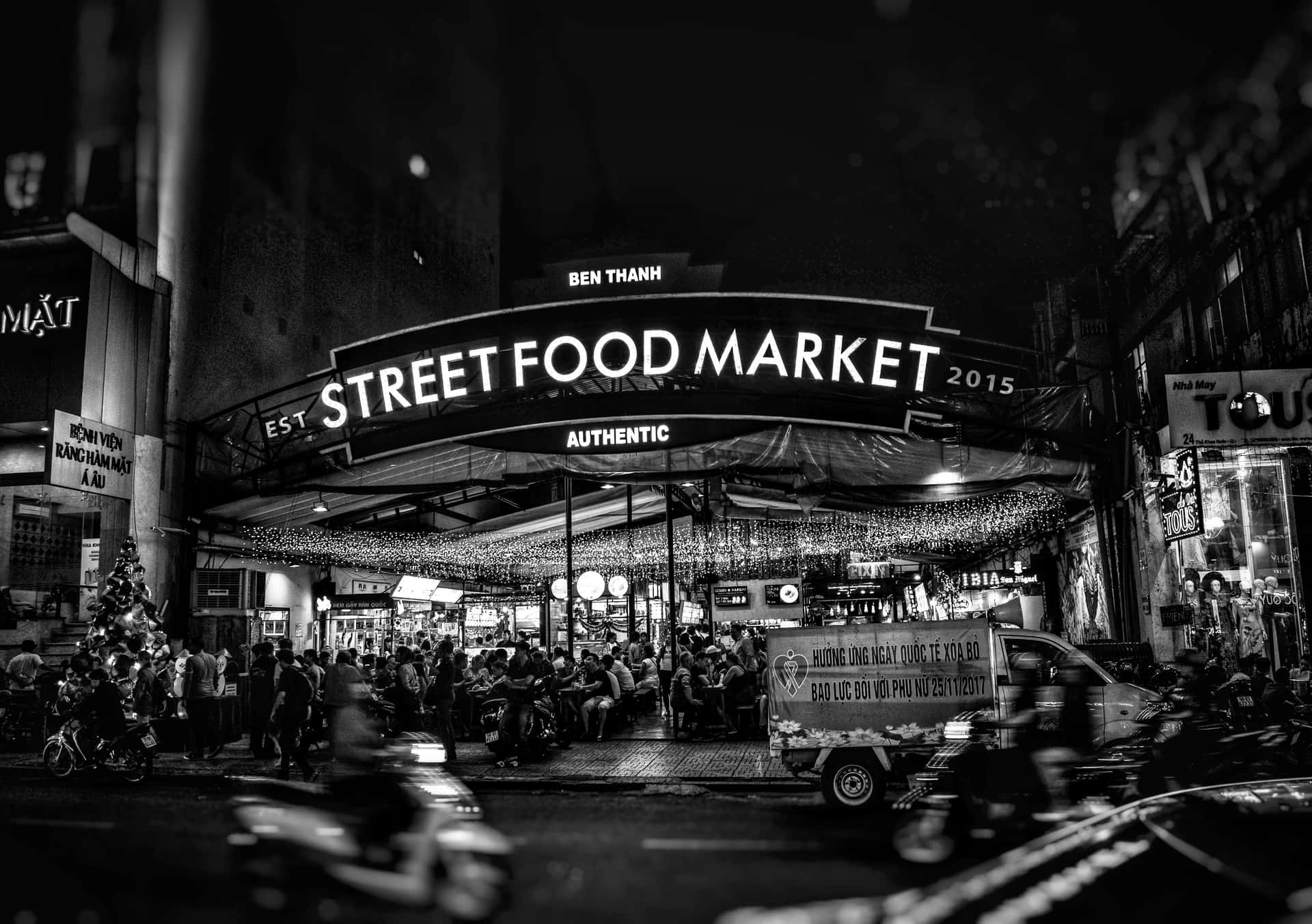  壁紙 — 濱城街頭美食市場熱鬧的黑白夜景，人群在發光招牌下用餐，電單車穿梭而過。