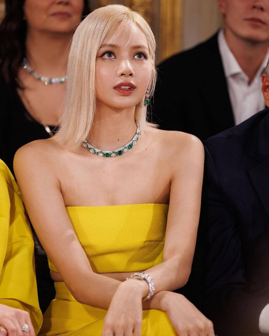 Bvlgari珠寶秀Lisa BLACKPINK高清照 壁紙 — 一位擁有鉑金色頭髮的女性穿著鮮黃色無肩帶連衣裙和翡翠珠寶，坐在優雅的活動中。