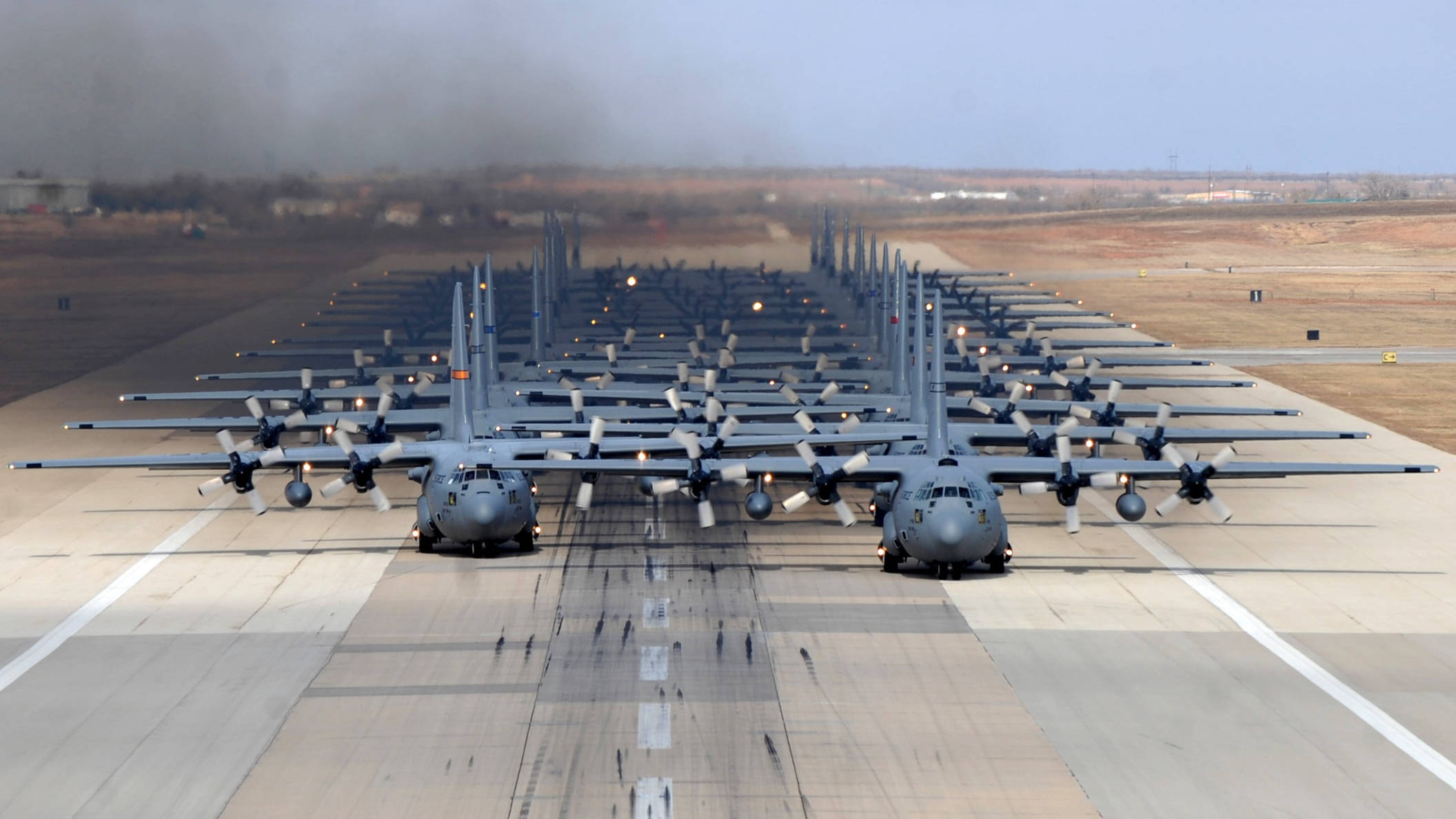 洛克希德C-130運輸機排列跑道 壁紙 — 一排灰色軍用運輸機排列在跑道上，天空朦朧，遠處有煙霧。