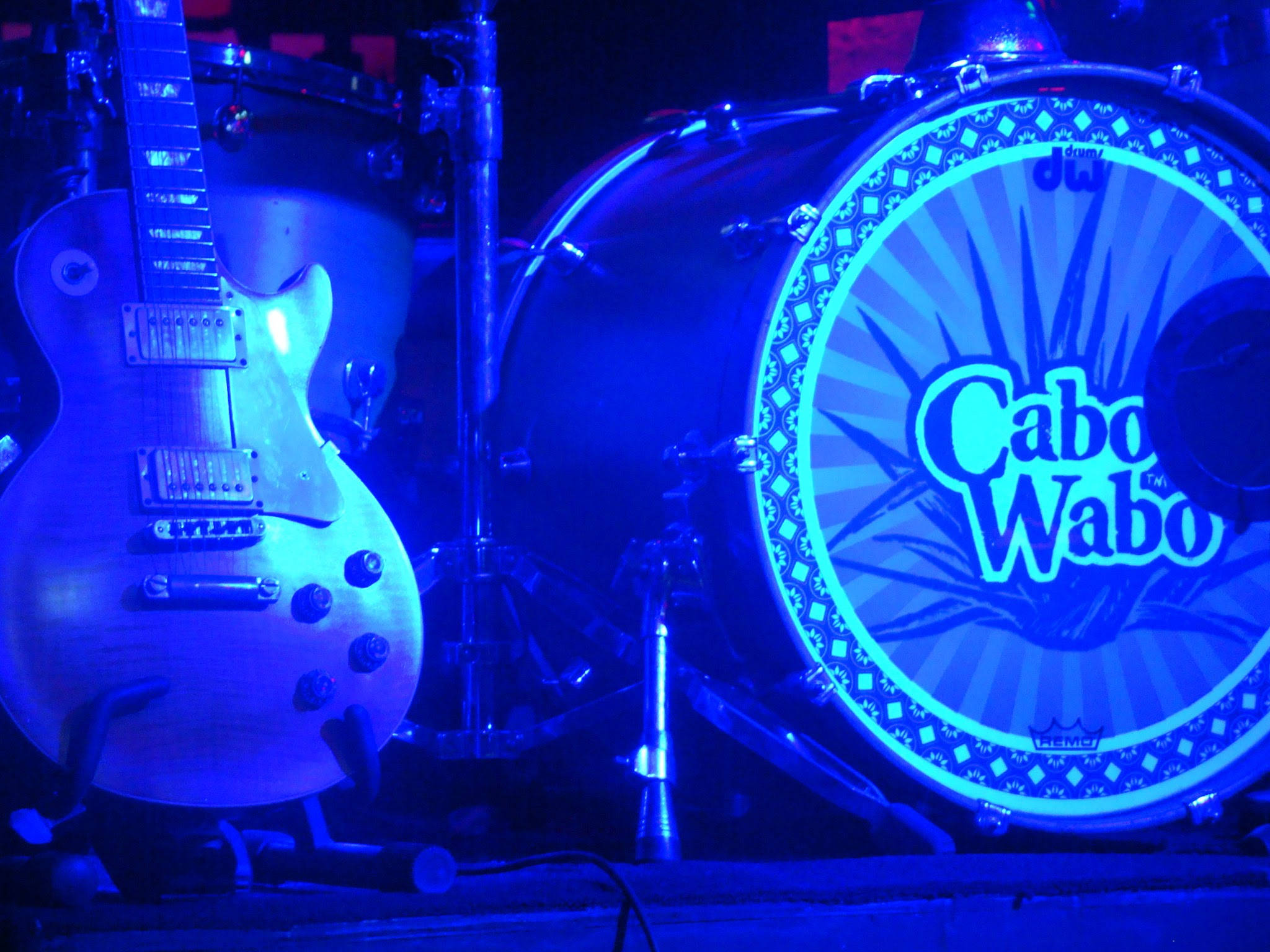 Cabo Wabo現場樂隊 壁紙 — 一支復古電結他和一個印有 'Cabo Wabo' 標誌的低音鼓，沐浴在強烈的藍色舞台燈光中。