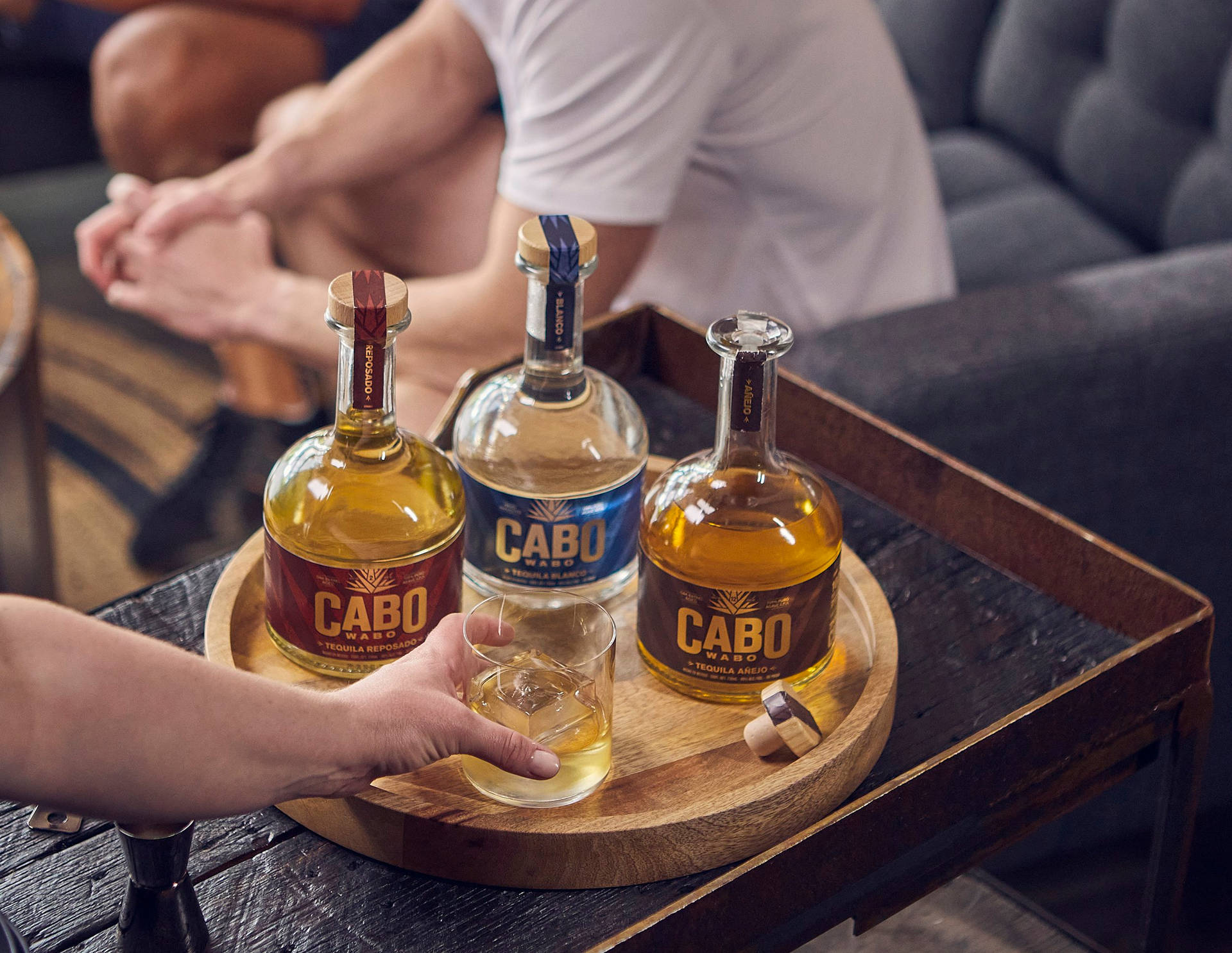 手將龍舌蘭酒倒入木托盤上的玻璃杯，旁邊有三支Cabo Wabo酒瓶。