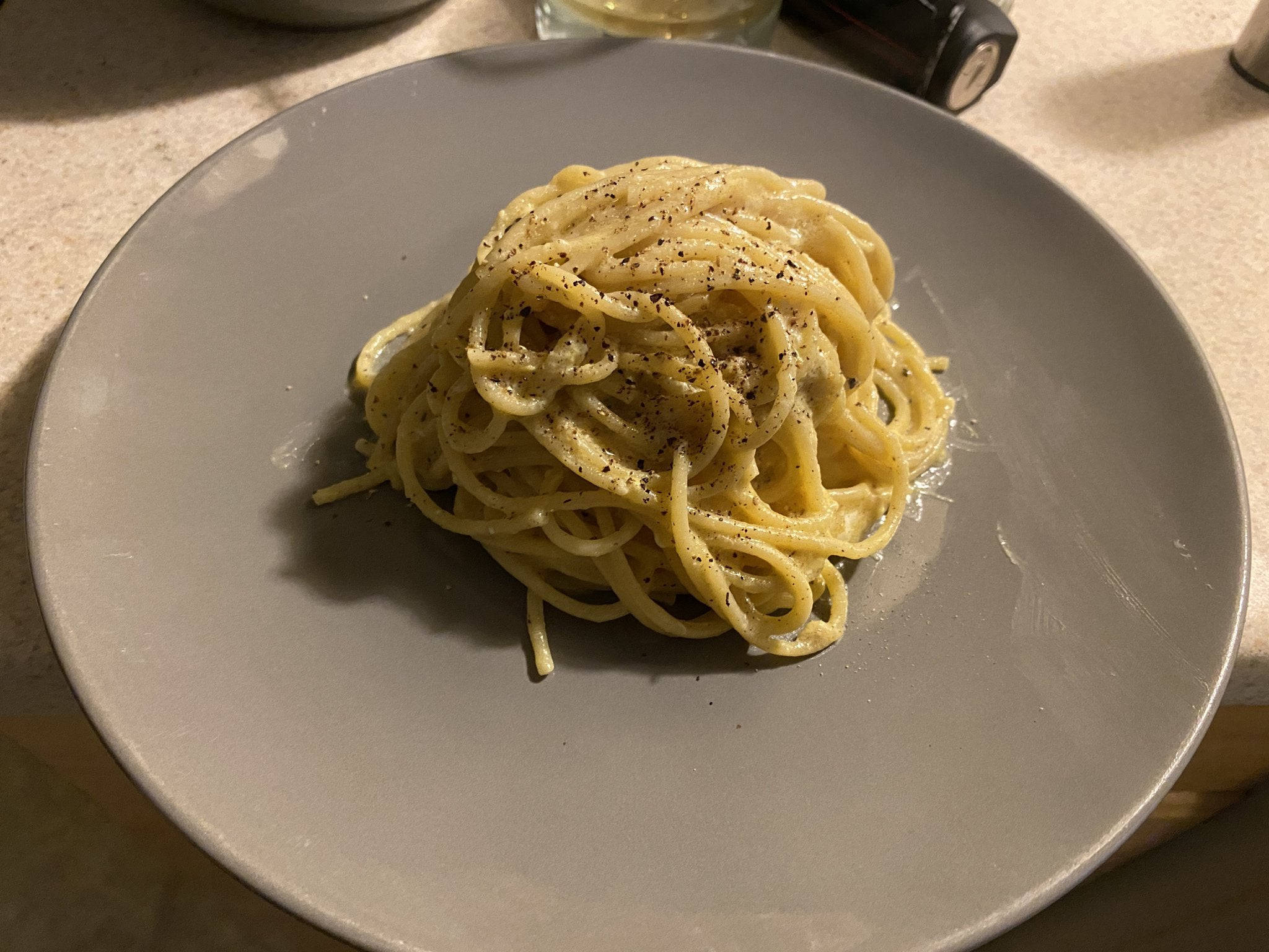 灰色盤盛裝小份 Cacio E Pepe 壁紙 — 一團奶油意粉配黑胡椒，在溫暖的室內燈光下置於灰色碟中央。