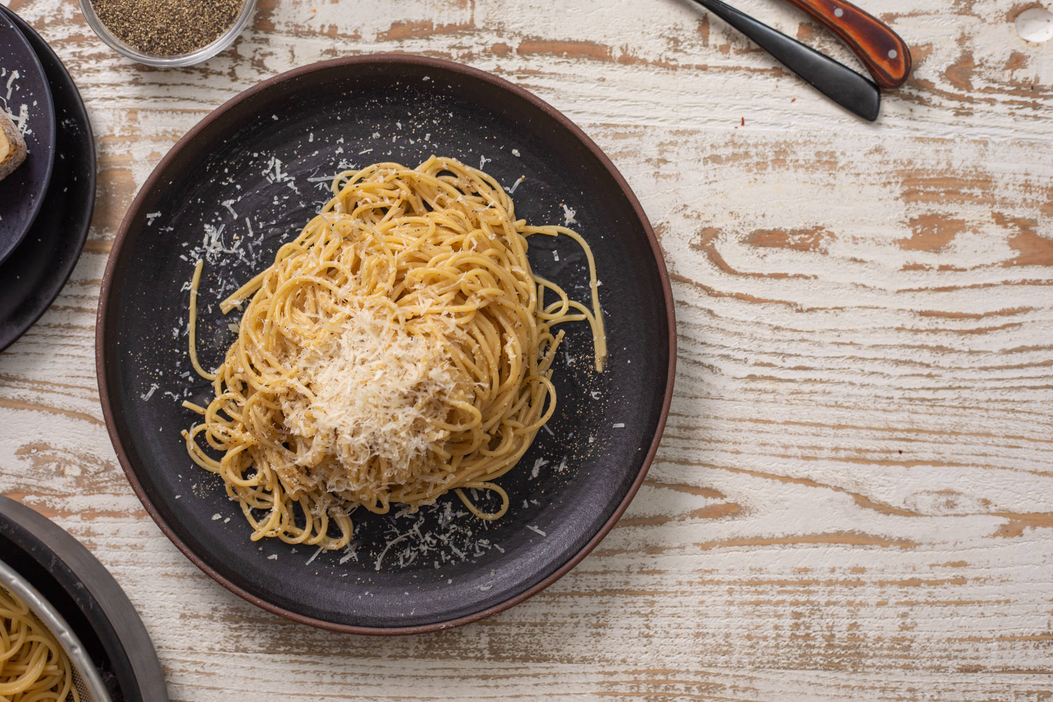 Cacio E Pepe 瓷器盤盛裝 壁紙 — 一盤鄉村風格的意大利麵，上面撒滿了磨碎的芝士，放在一張風化的木桌上，附近有廚房工具。
