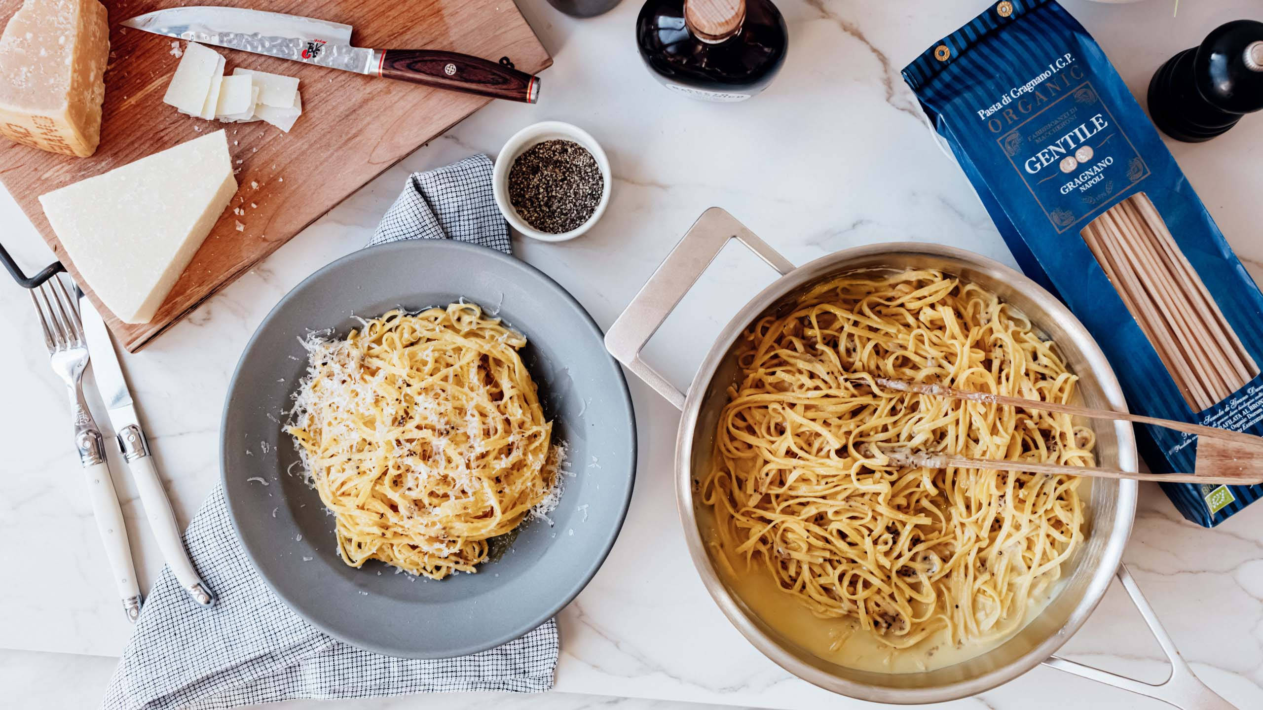 Cacio E Pepe 食材 壁紙 — 從上方俯瞰新鮮烹煮的意大利麵在鍋中，旁邊是撒上芝士碎的碟上菜餚，並被巴馬臣芝士和黑胡椒等食材包圍。