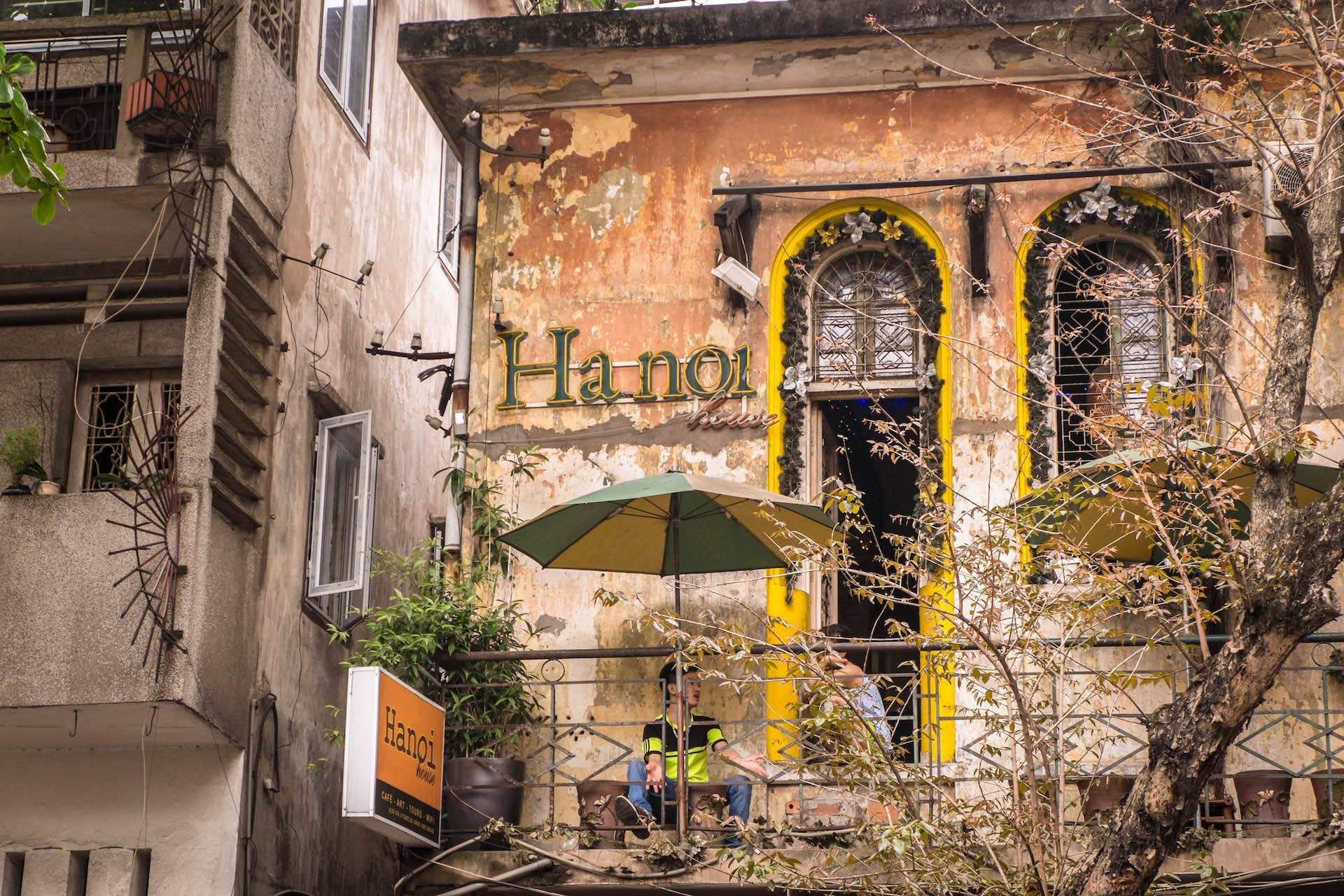 河內Café House咖啡館 壁紙 — 一座褪色建築，橙色油漆剝落，黃色拱形窗戶，內有一間咖啡館，顧客坐在綠色遮陽傘下。