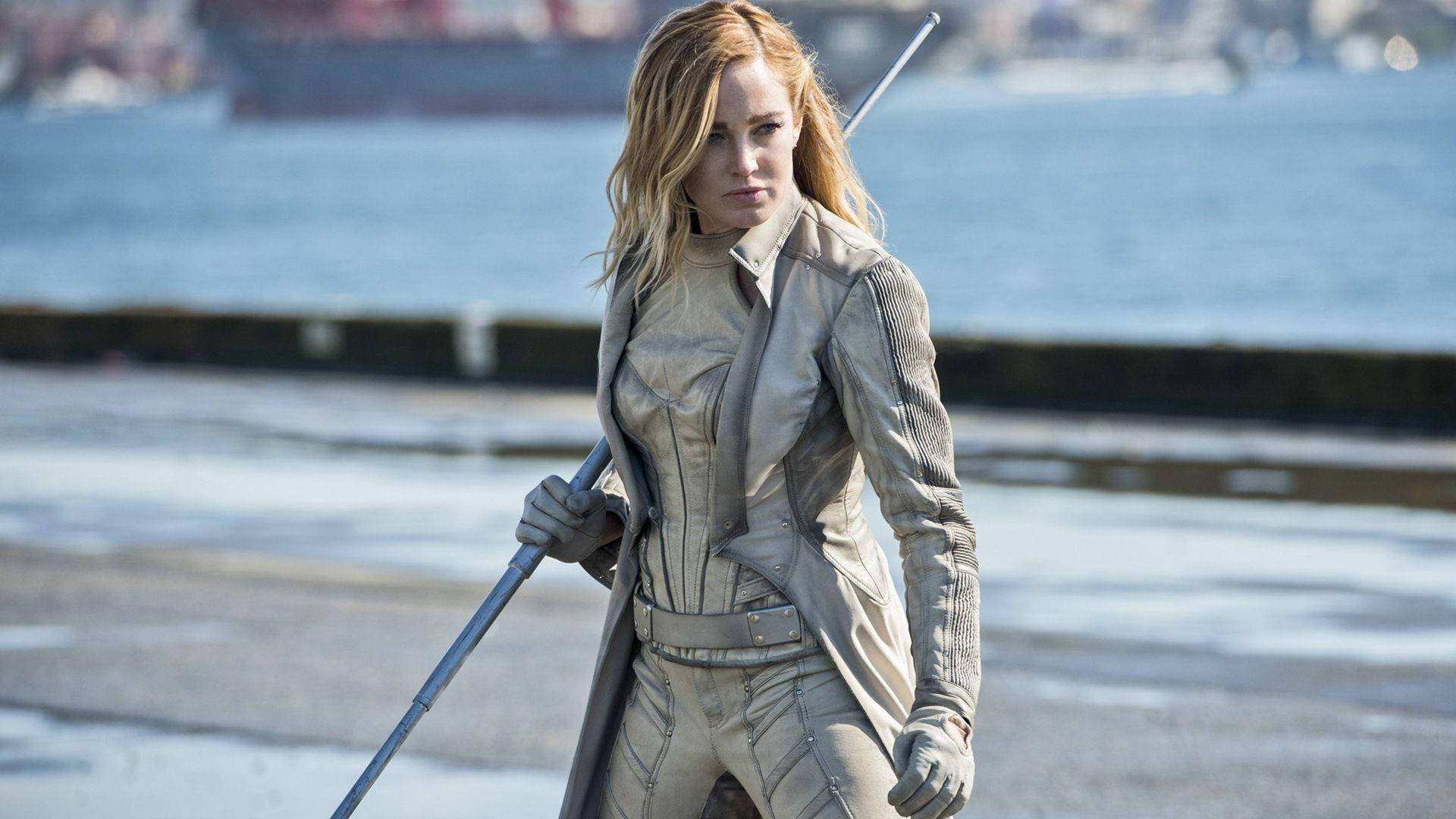 Caity Lotz 飾白雀鴉 壁紙 — 一位金髮女子身穿米色戰術服，手持法杖，站在碼頭上，背景有一艘船。