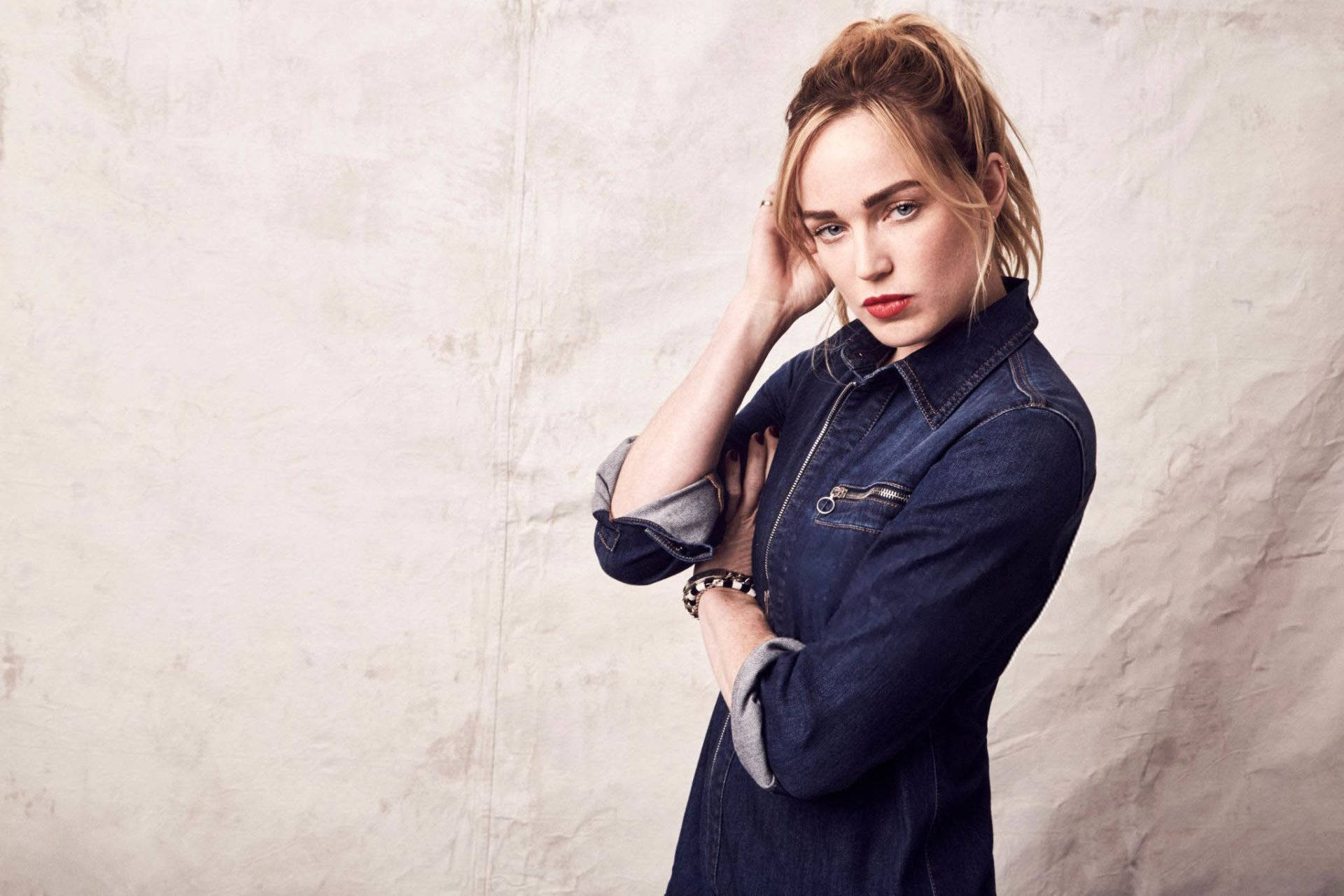 Caity Lotz 藍色牛仔裝 壁紙 — 一位身穿牛仔外套的女性，在帶有紋理的米色牆壁前自信地擺姿勢，一隻手靠近臉部。