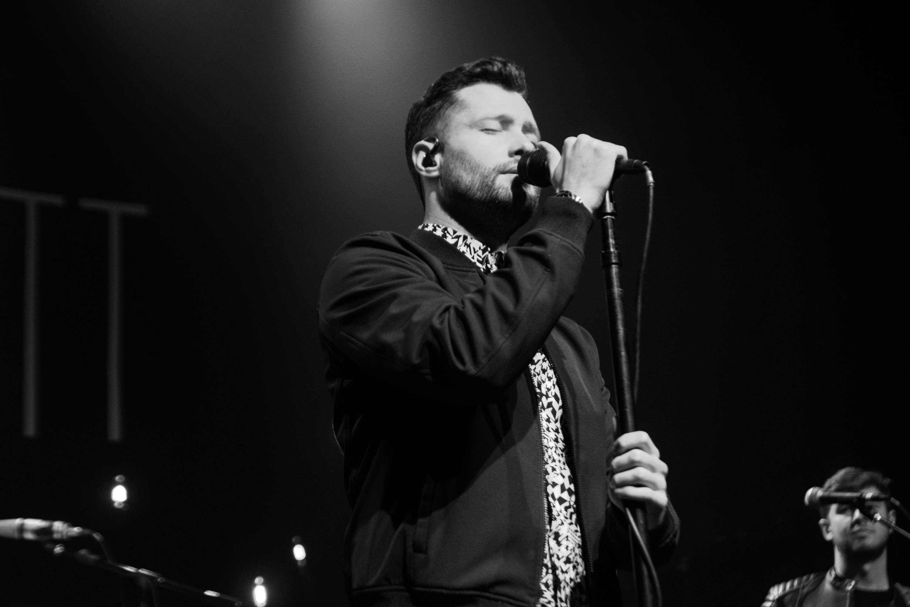 Calum Scott 舞台演唱 壁紙 — 一張黑白照片，顯示一位男歌手在聚光燈下熱情地在舞台上表演，背景中可見一位樂隊成員。