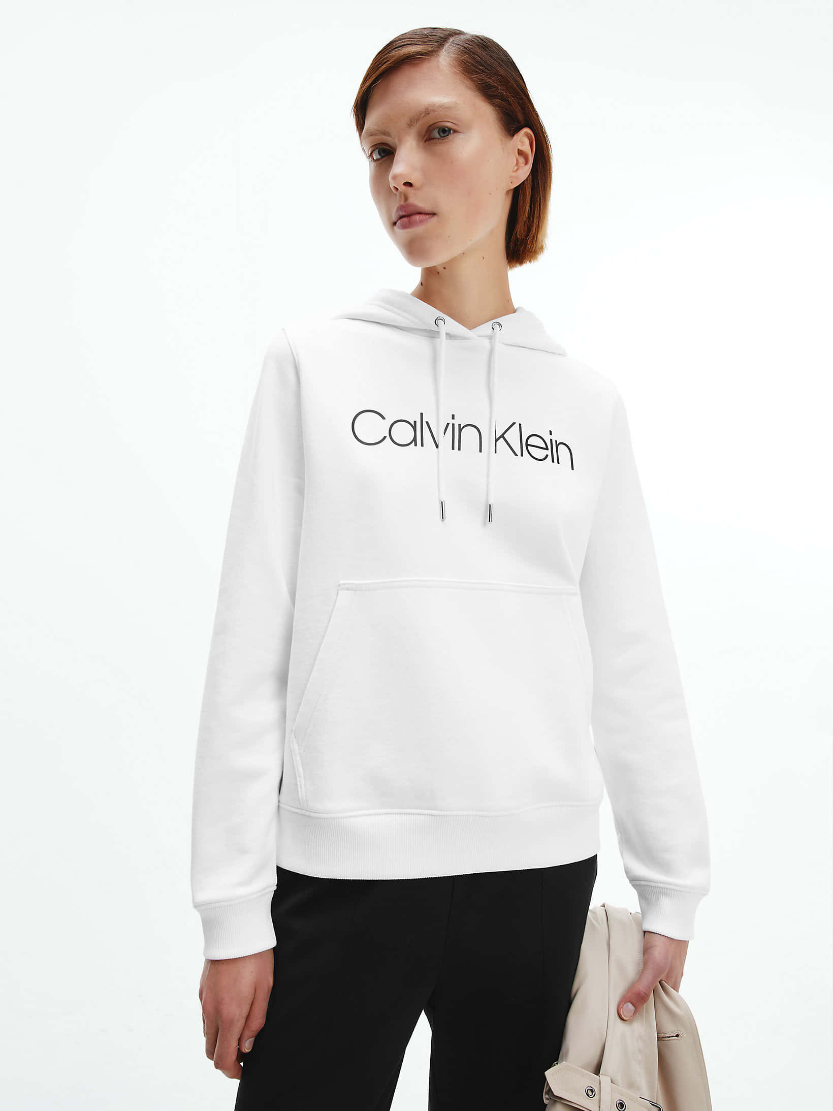 以全新Calvin Klein時尚脫穎而出 壁紙 — 一位身穿白色Calvin Klein連帽衫和黑色褲子的女性，在純白色背景前拿著一個米色手袋。