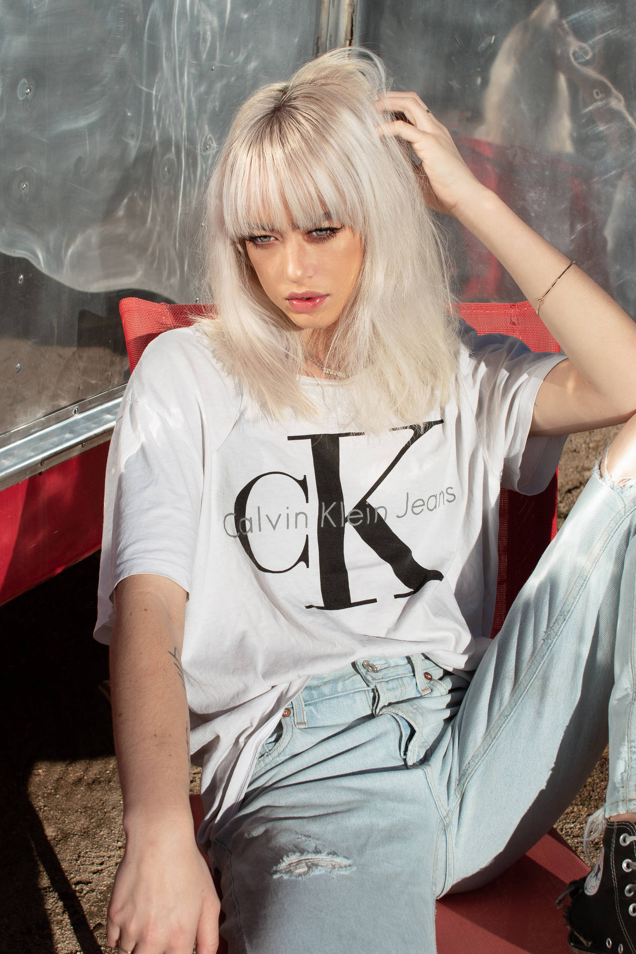 年輕女性留著金色頭髮，穿著Calvin Klein Jeans T恤和破洞牛仔褲，坐在紅色椅子上。