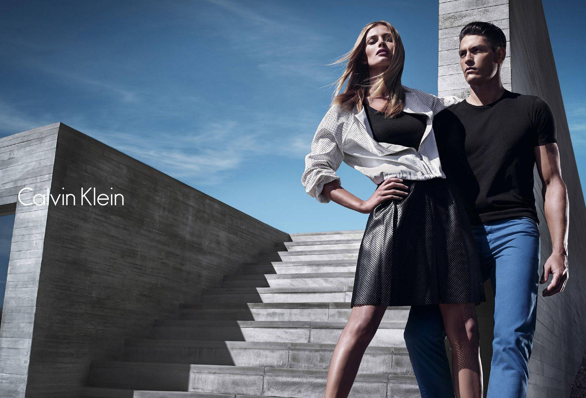 穿著 Calvin Klein 服裝的一男一女站在藍天下的混凝土樓梯上。