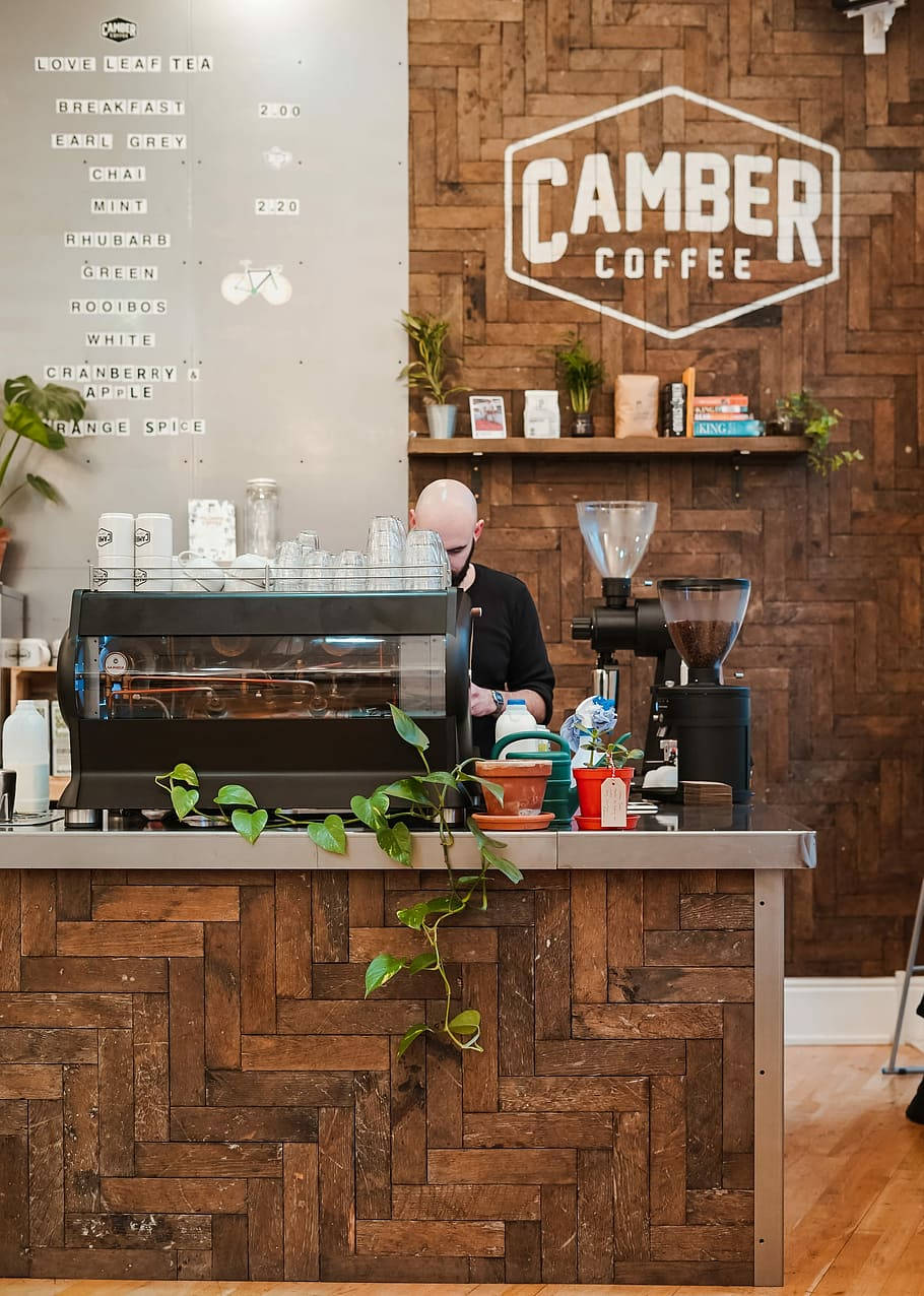 Camber 咖啡店 壁紙 — 一位身穿黑色襯衫的咖啡師在Camber Coffee的木製人字形櫃檯後準備咖啡，旁邊有菜單板和綠色植物。