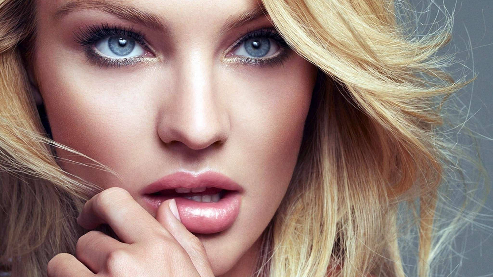 Candice Swanepoel 近距離特寫攝影 壁紙 — 一位金髮女性的特寫肖像，擁有銳利的藍色眼睛和光澤的嘴唇，她的手輕輕觸碰嘴巴，背景為柔和的灰色。