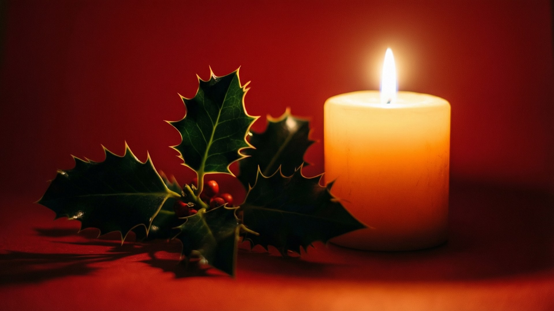 一支點燃的蠟燭旁邊放著帶有紅色漿果的冬青枝，在紅色表面上