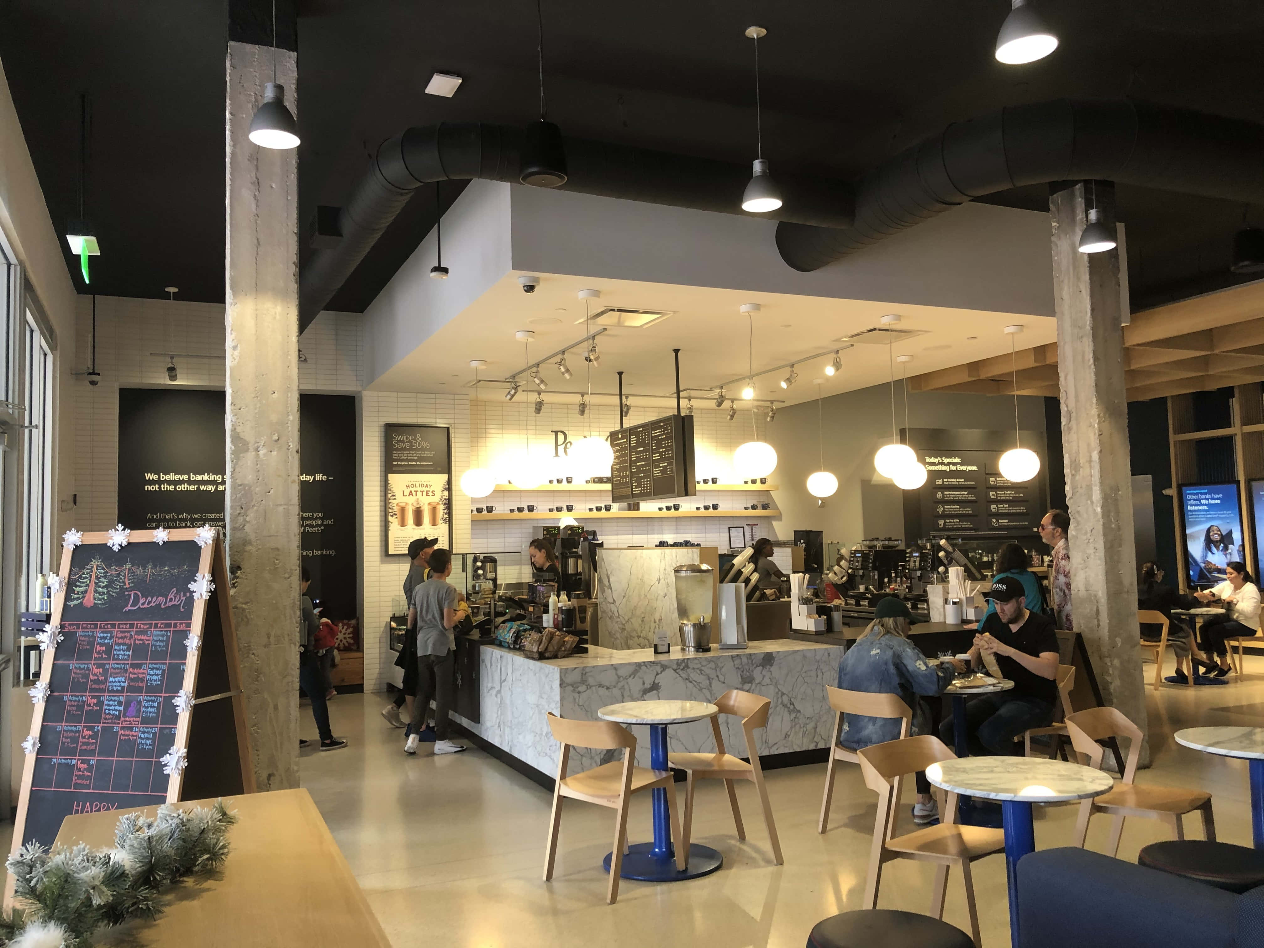 Capital One咖啡廳入口 壁紙 — 現代咖啡店，設有外露管道、木樑、大理石檯面，顧客在明亮通風的空間中享用飲品。