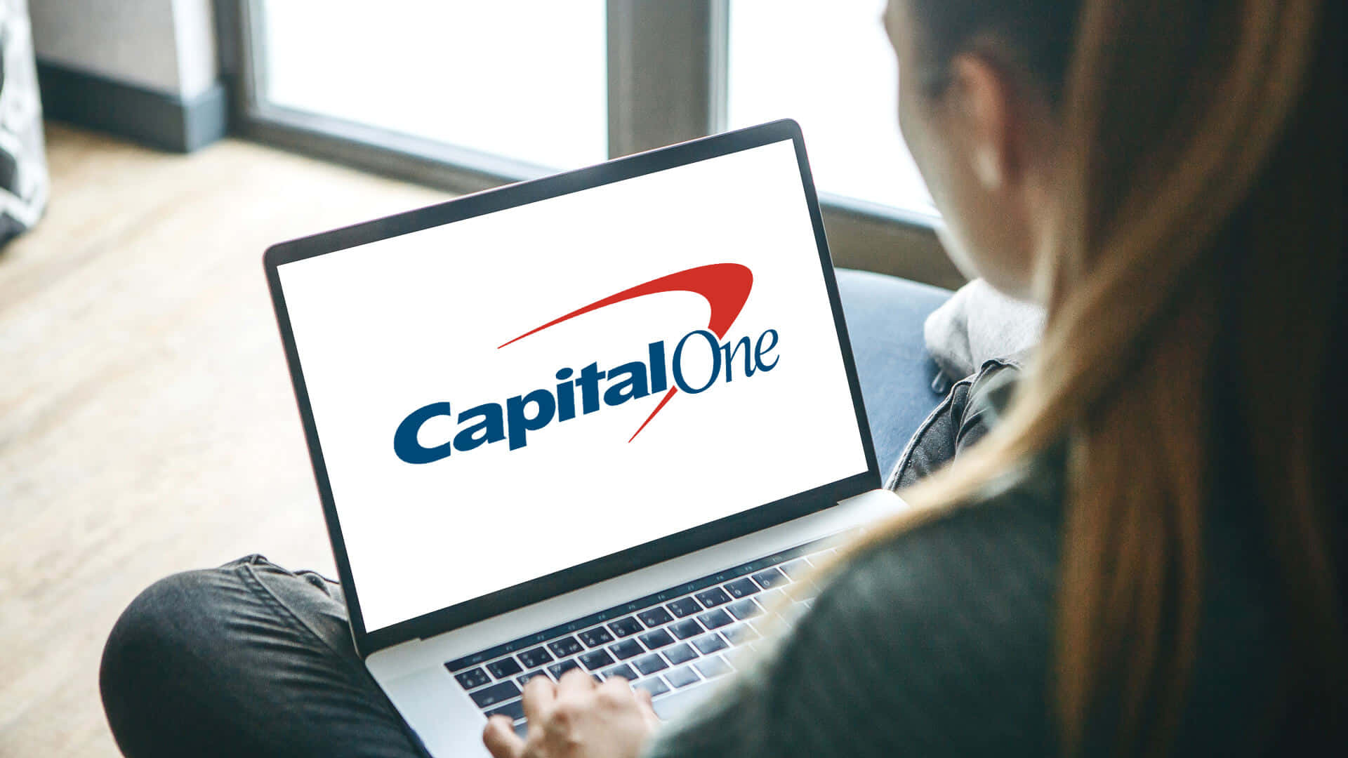 筆記型電腦螢幕顯示 Capital One 標誌 壁紙 — 一個人盤腿坐在沙發上，在窗邊使用一部顯示Capital One標誌、屏幕明亮的筆記型電腦。