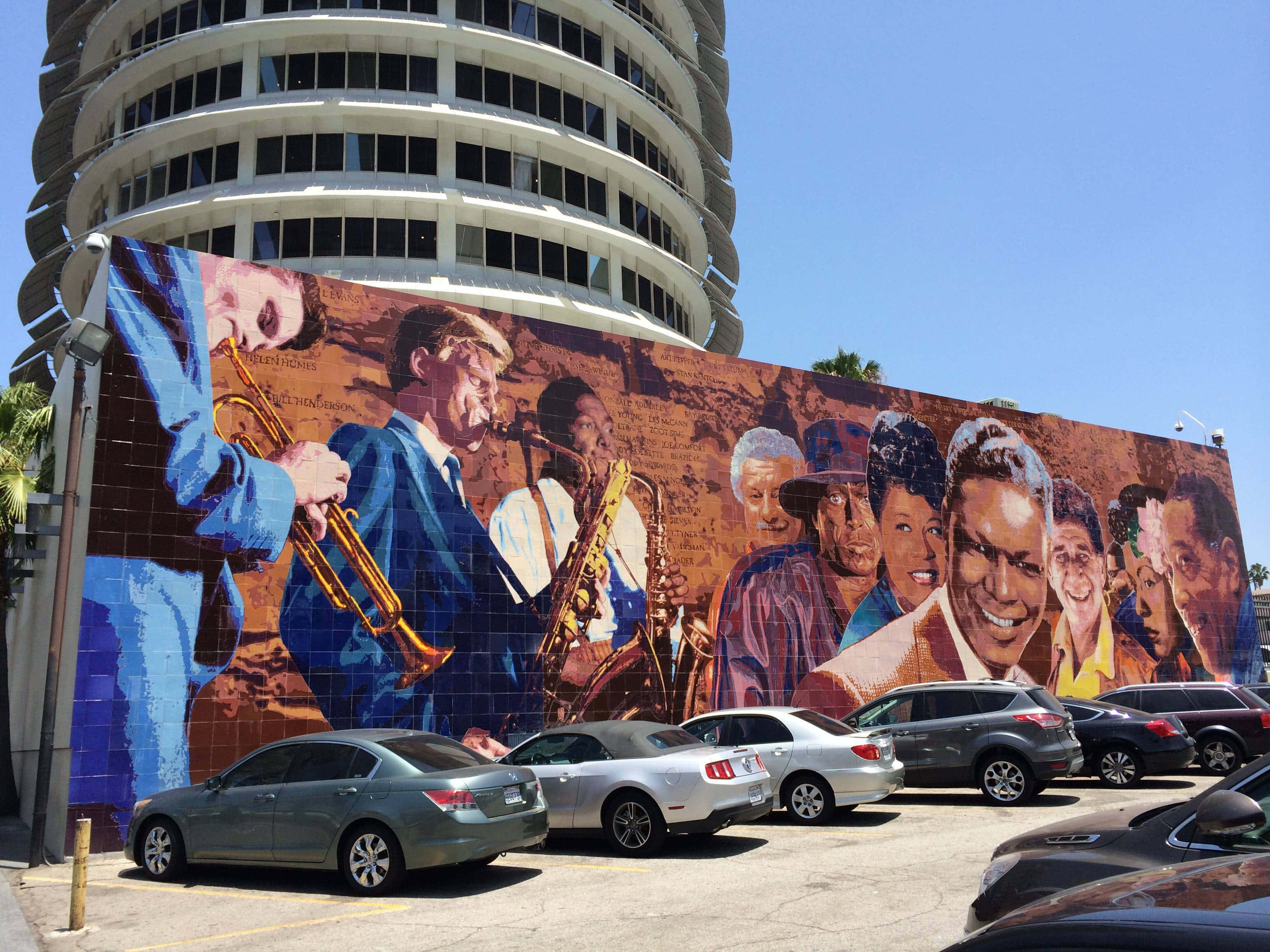 Capitol Records大樓地面層的壁畫 壁紙 — 在晴朗的藍天下，一座弧形建築物的側面裝飾著一幅充滿活力的爵士樂手和笑臉壁畫。