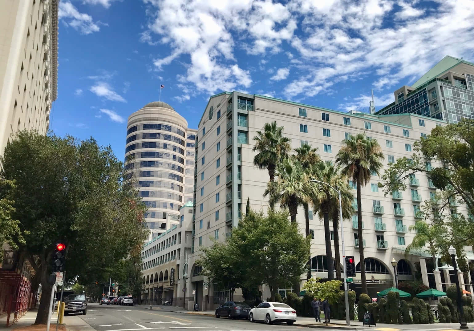 Capitol Records大樓的遠景 壁紙 — 一條陽光明媚的城市街道，兩旁種滿棕櫚樹和現代建築，頭頂是明亮的藍天，白雲點綴其間。