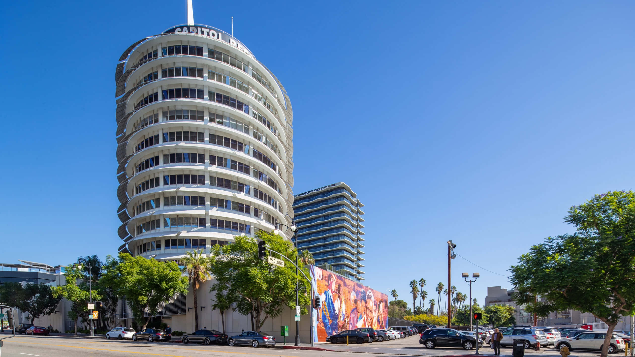 Capitol Records大樓的特寫 壁紙 — 一座引人注目的白色弧形 Capitol Records 大樓聳立在陽光明媚、種滿棕櫚樹和停滿汽車的街道上。