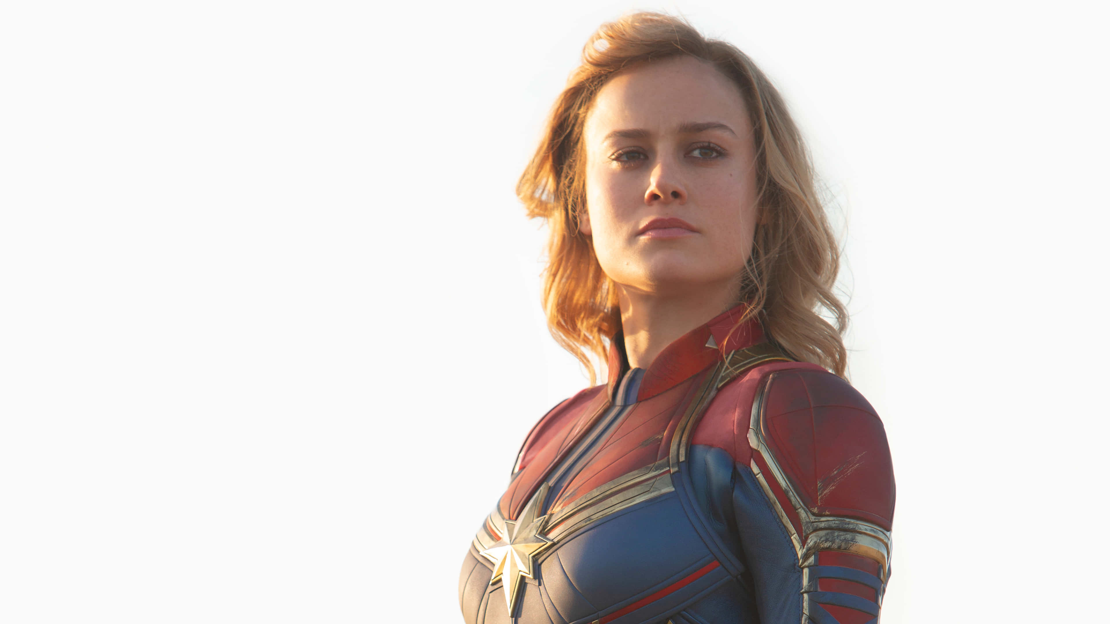 brie larson 桌布 壁紙 — Captain Marvel 側身站在明亮的白色背景前，她的紅藍金色套裝在柔和光線下閃閃發光。