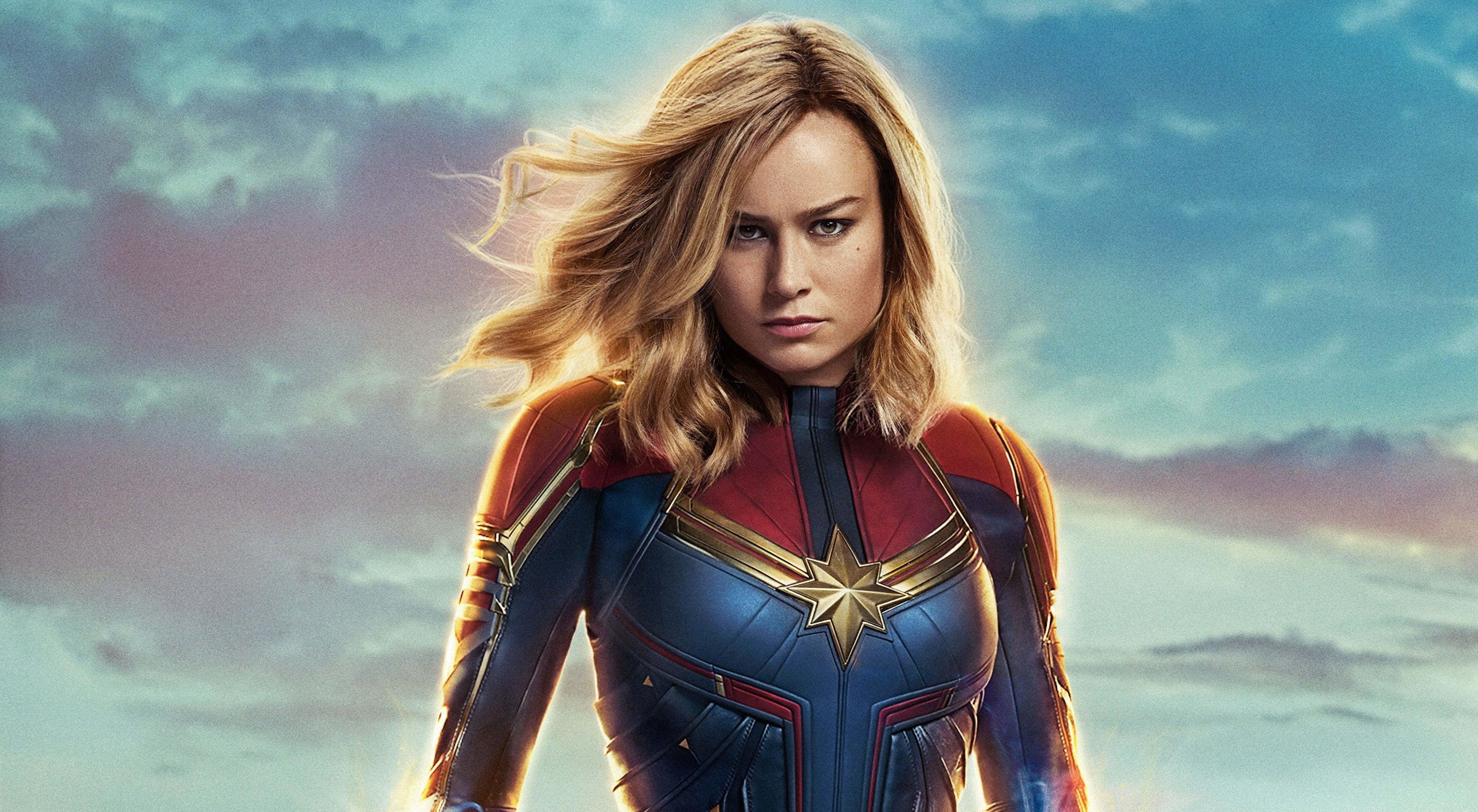 4K Marvel 壁紙 桌布 壁紙 — Captain Marvel 自信地穿著她的藍色和紅色套裝，站在戲劇性的暮色天空下，金髮隨風飄揚。