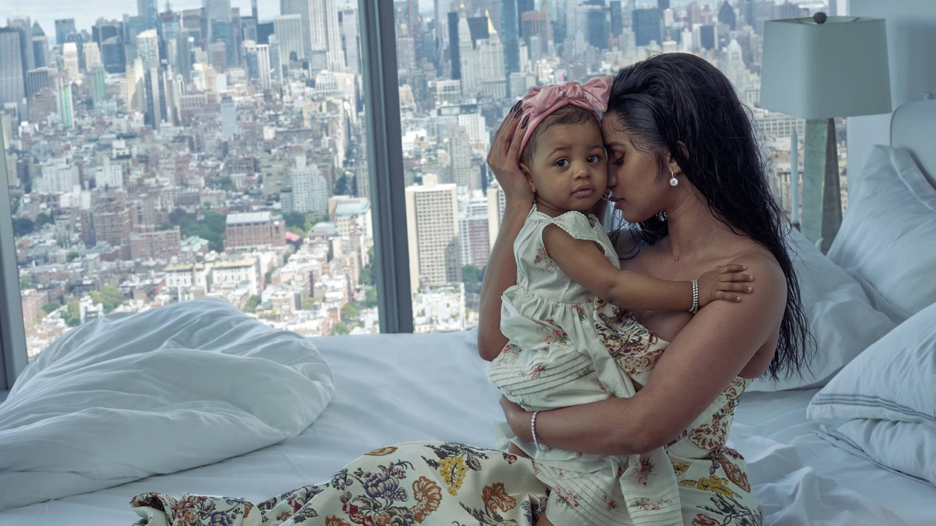 Cardi B 與 Kulture 壁紙 — 母親在床上一邊透過大窗戶欣賞密集的城市天際線全景，一邊溫柔地親吻她的女嬰。