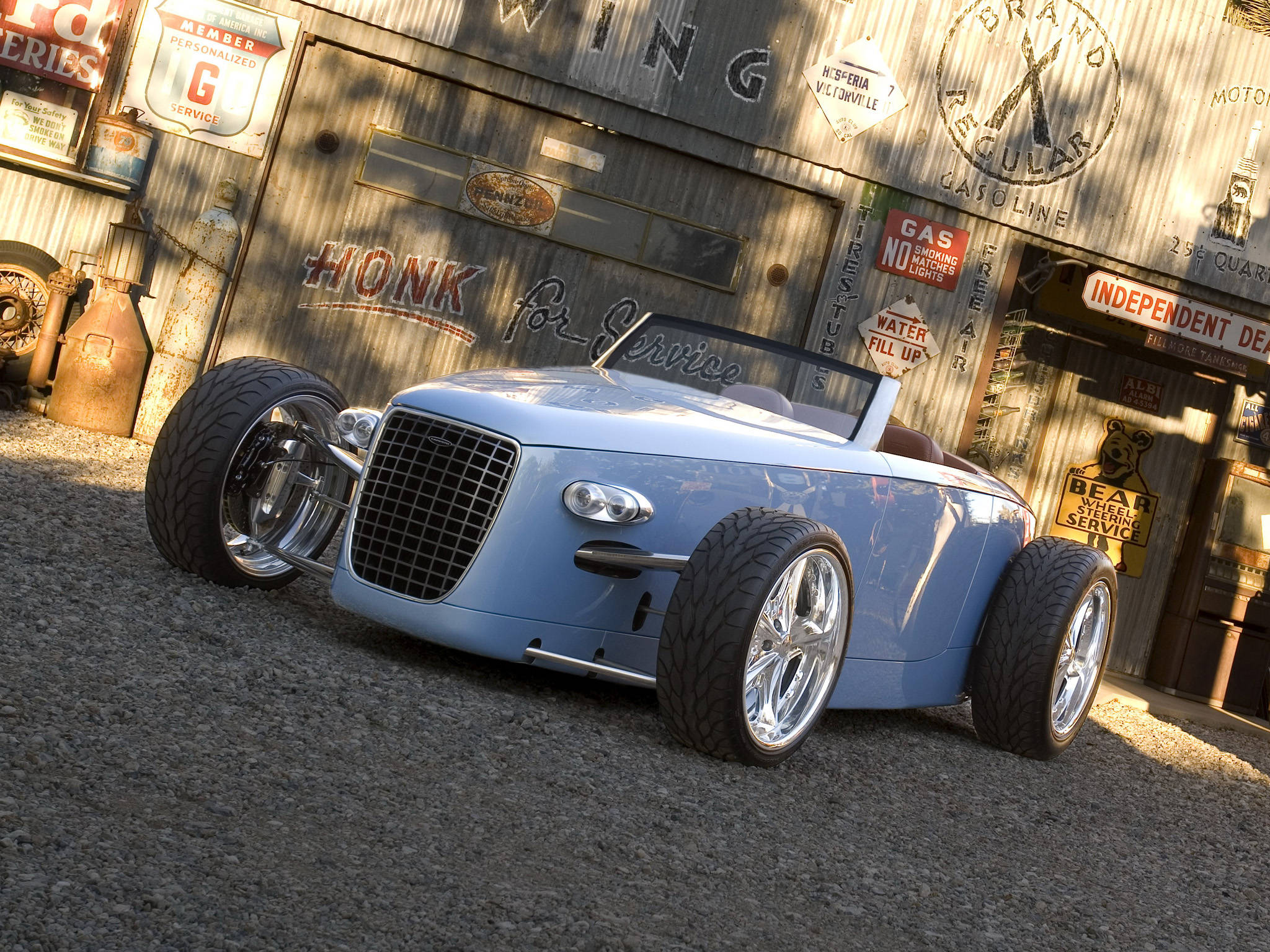 Caresto Speedster Volvo iPhone 壁紙 — 一輛淺藍色定制熱棒車配備鍍鉻輪圈，停在一個裝飾著復古標誌、飽經風霜的復古加油站前。