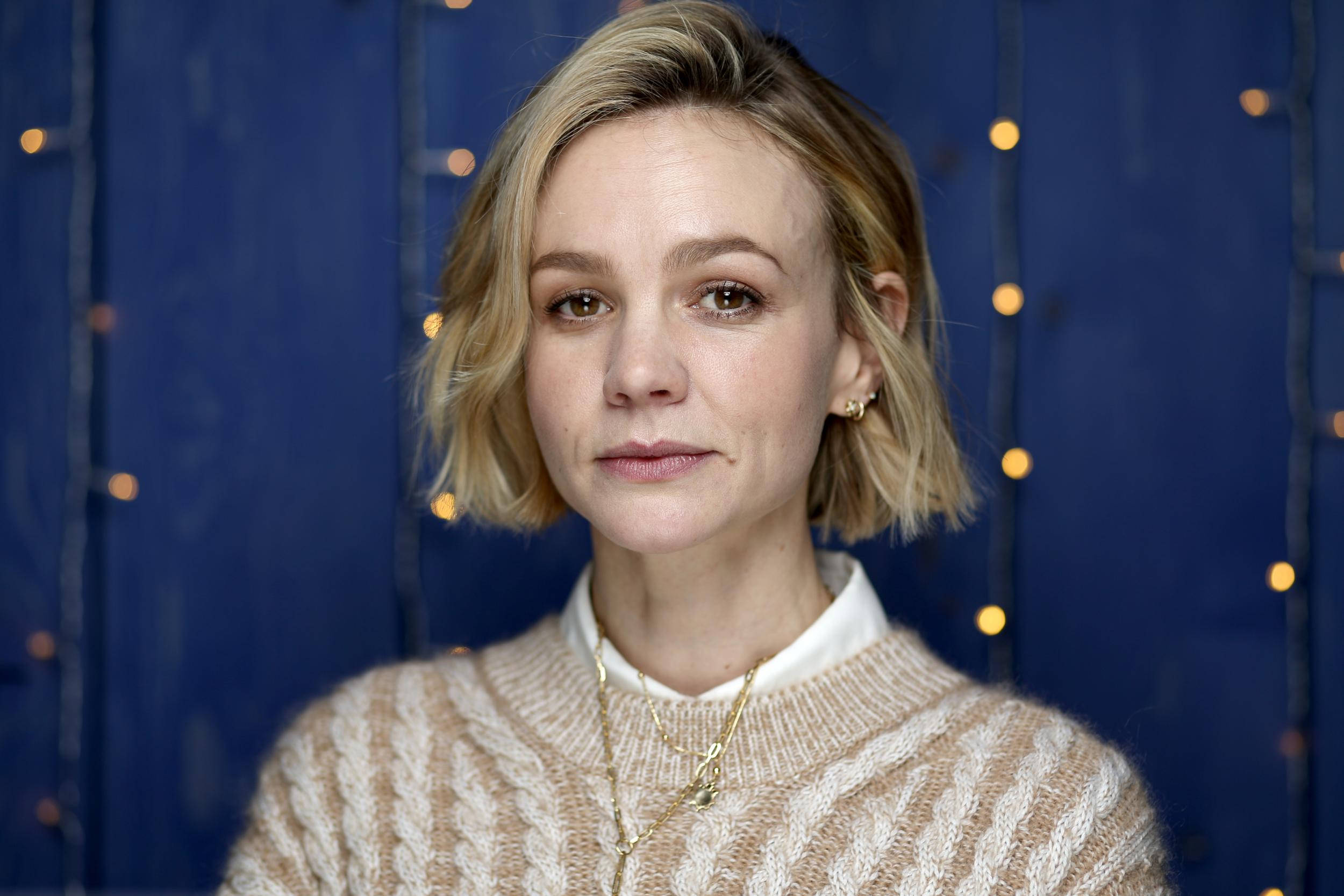 Carey Mulligan穿毛衣 壁紙 — 一位短金髮女性穿著米色電纜針織毛衣，背景是藍色和溫暖的串燈。