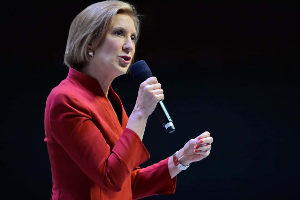 Carly Fiorina 手勢表達 壁紙 — 一位身穿紅色西裝外套的女性在黑暗背景和聚焦燈光下對著麥克風說話。