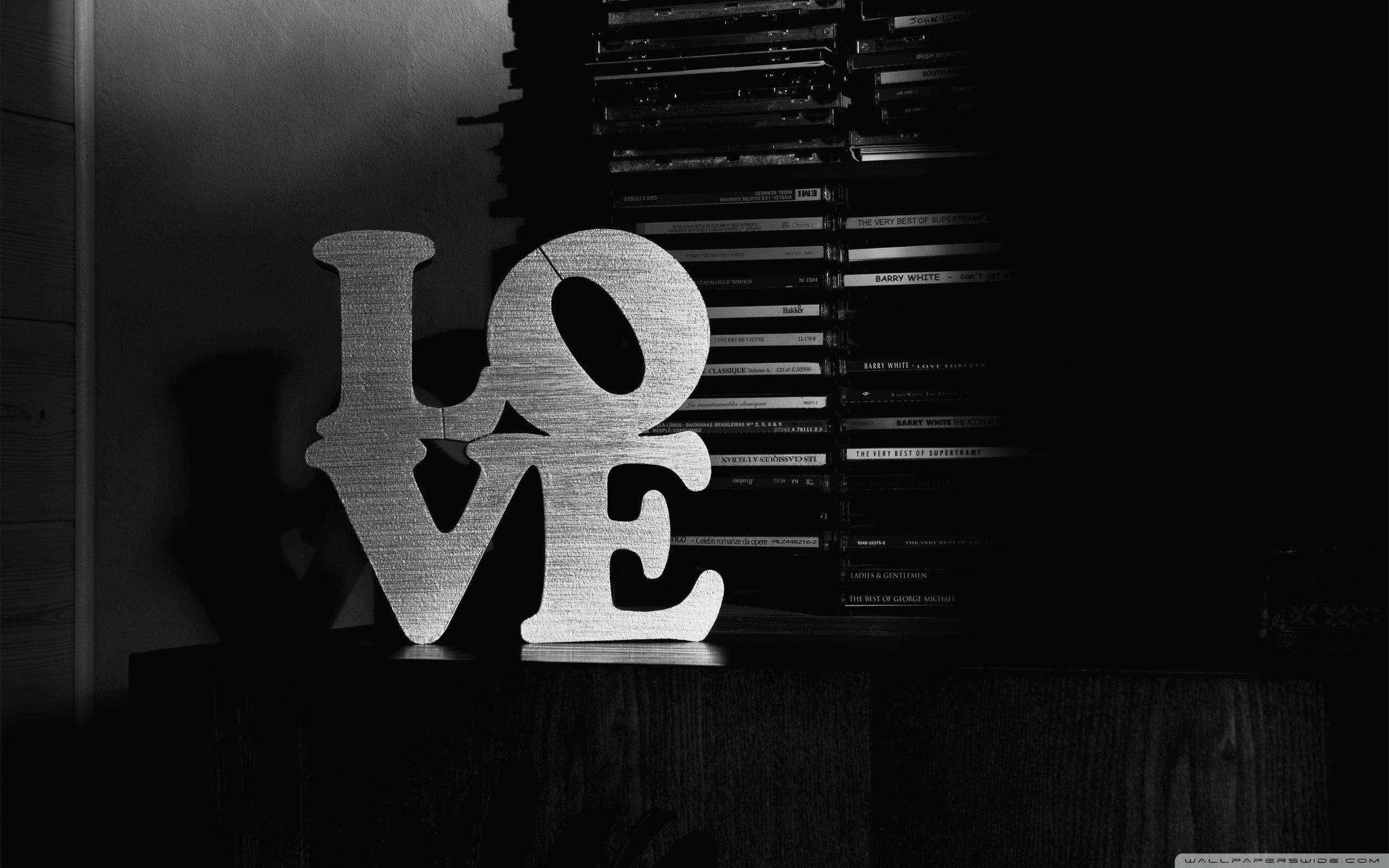 黑愛 字母 壁紙 — 在昏暗的房間裡，一個單色的「LOVE」標誌在木架上柔和地發光，旁邊是一疊高聳的DVD。
