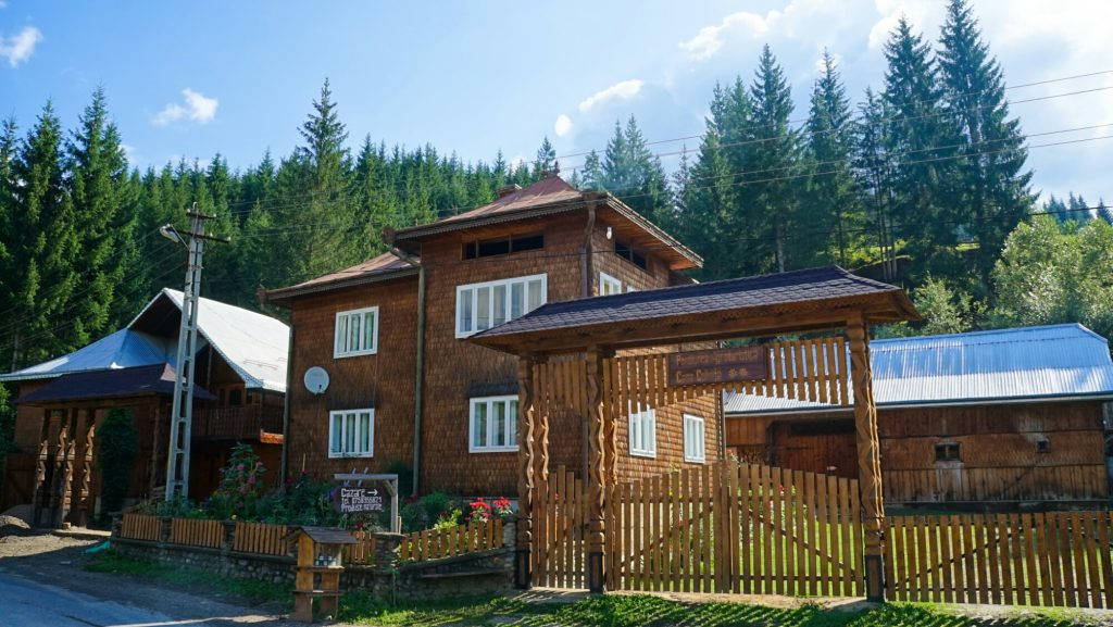 羅馬尼亞 Casa Colinița 農莊 壁紙 — 一座帶有門的質樸木屋矗立在陽光明媚的森林空地上，頭頂是明亮的藍天。