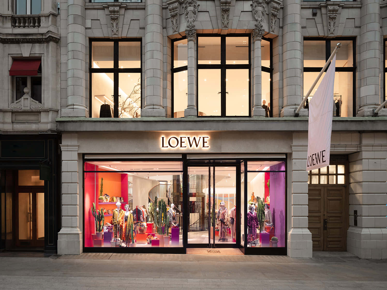 倫敦Casa Loewe外觀 壁紙 — 一間宏偉的Loewe精品店，擁有華麗的石頭建築和充滿活力的粉紫色室內燈光，照亮了人體模型和仙人掌展示。