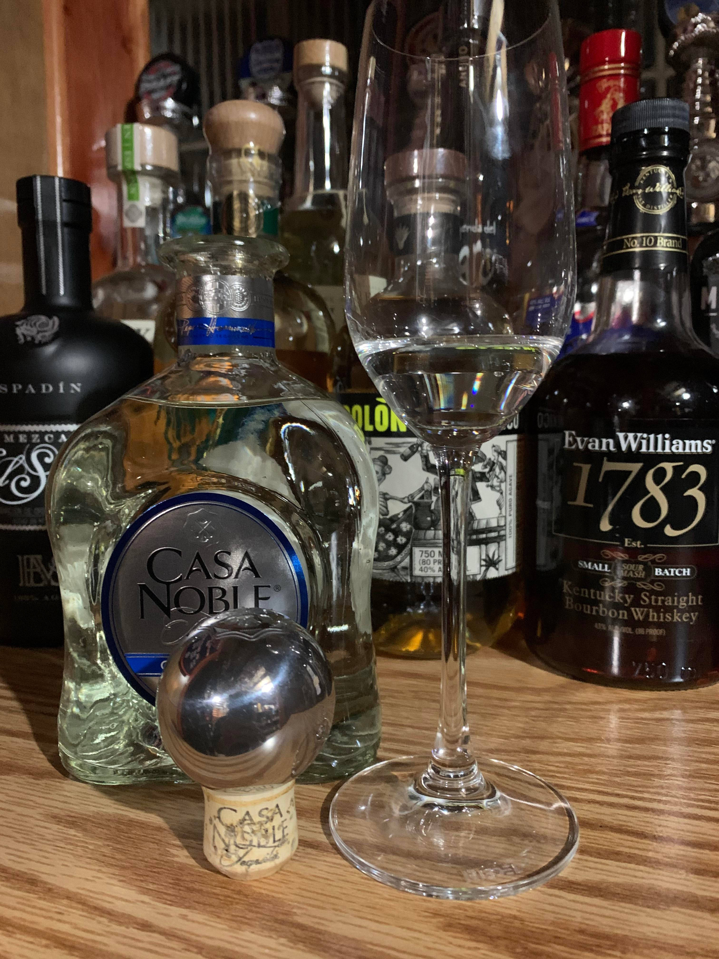 Casa Noble Crystal Blanco 龍舌蘭酒盛於香檳杯 壁紙 — Casa Noble 龍舌蘭酒瓶、酒杯和開瓶器的特寫，背景是木製吧檯上還有其他酒瓶。