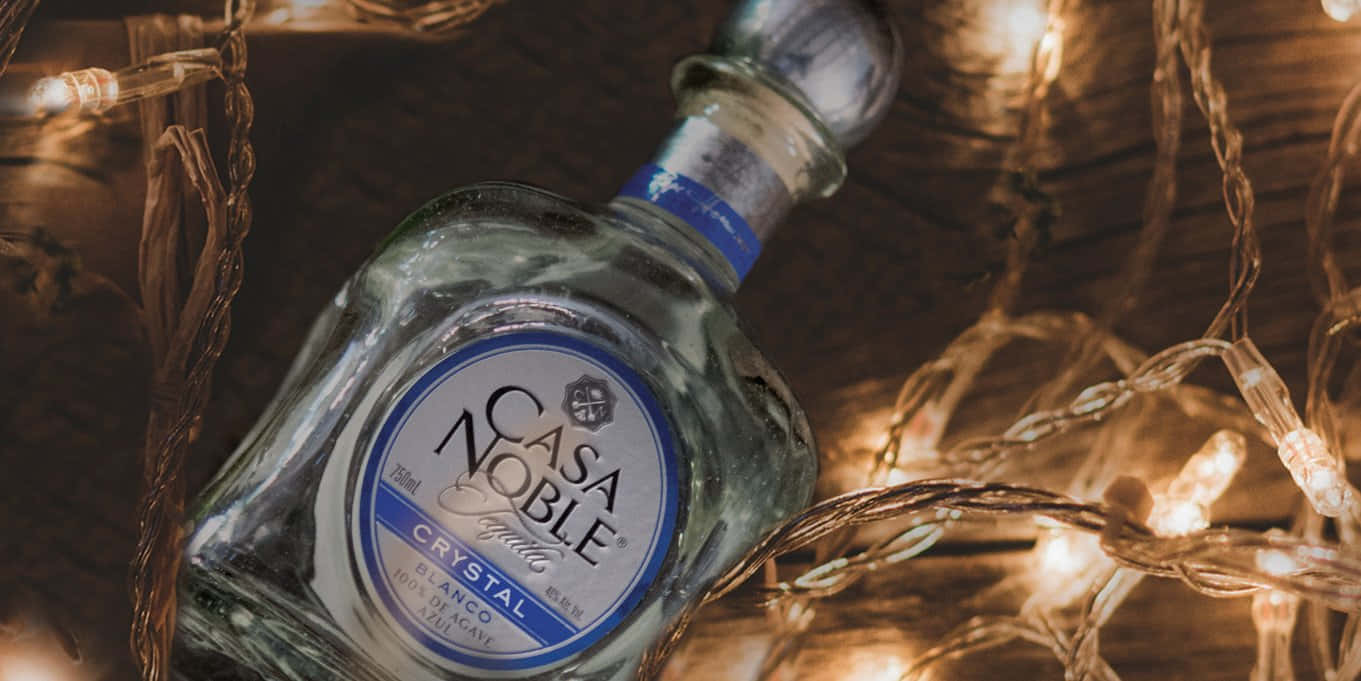 Casa Noble Crystal Blanco聖誕燈光 壁紙 — 一瓶透明的 Casa Noble Crystal Blanco 龍舌蘭酒放在木質表面上，周圍環繞著溫暖發光的串燈。