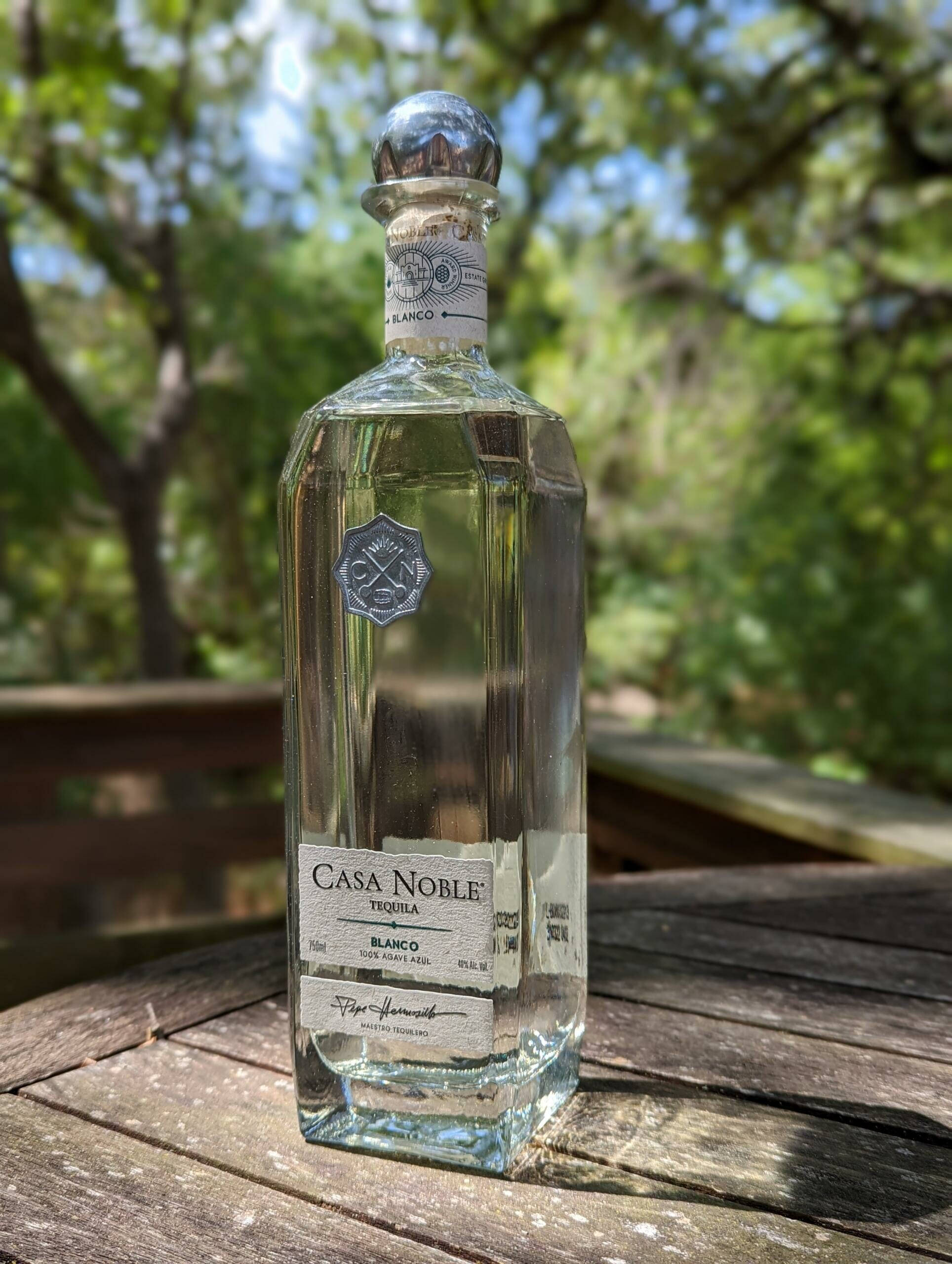 Casa Noble Nouvel白龍舌蘭，精緻蒸餾龍舌蘭 壁紙 — 一瓶透明的Casa Noble Blanco龍舌蘭酒放在戶外一張風化的木桌上，背景有柔焦的綠色樹木。