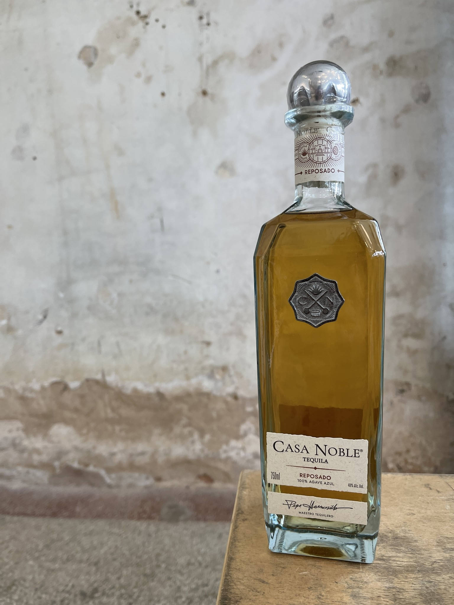 Casa Noble Reposado Nouvel有機龍舌蘭 壁紙 — 一個透明玻璃瓶的Casa Noble Reposado龍舌蘭酒，裝有琥珀色液體，放在木質表面上，背景是風化的混凝土牆。