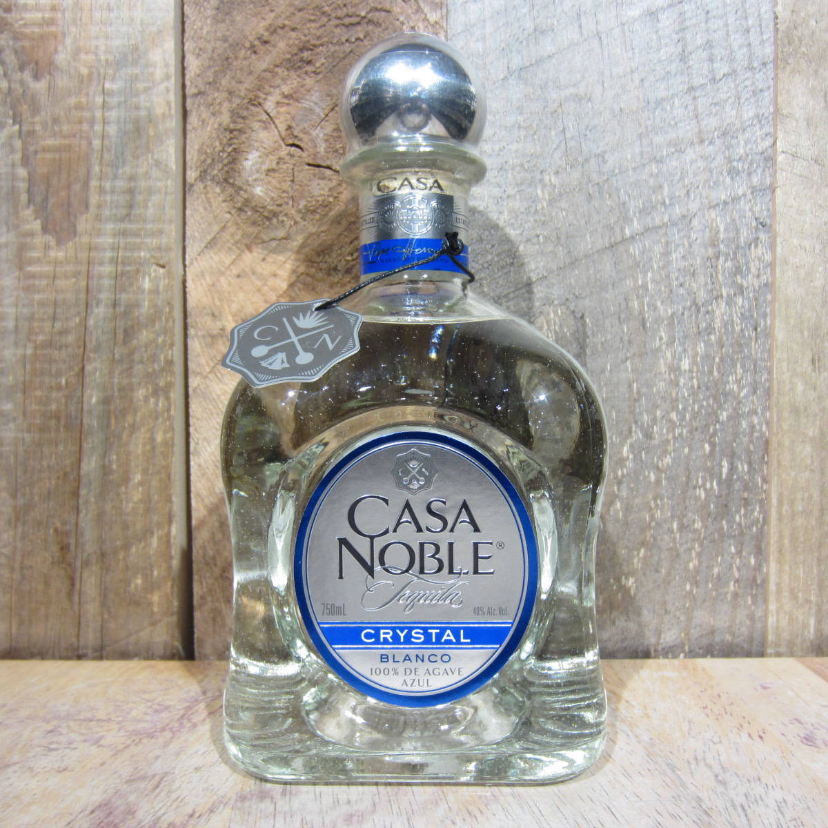 Casa Noble水晶白藍龍舌蘭 壁紙 — 一個透明的Casa Noble Crystal Blanco龍舌蘭酒瓶，帶有銀色瓶蓋和藍色標籤，放置在木質表面上，背景是質樸的木頭。
