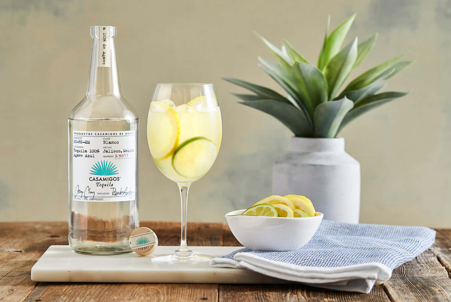 Casamigos 白色龍舌蘭酒搭配檸檬青檸氣泡水 壁紙 — 一瓶Casamigos龍舌蘭酒放在大理石托盤上，旁邊有一杯柑橘雞尾酒、盆栽植物和青檸片。